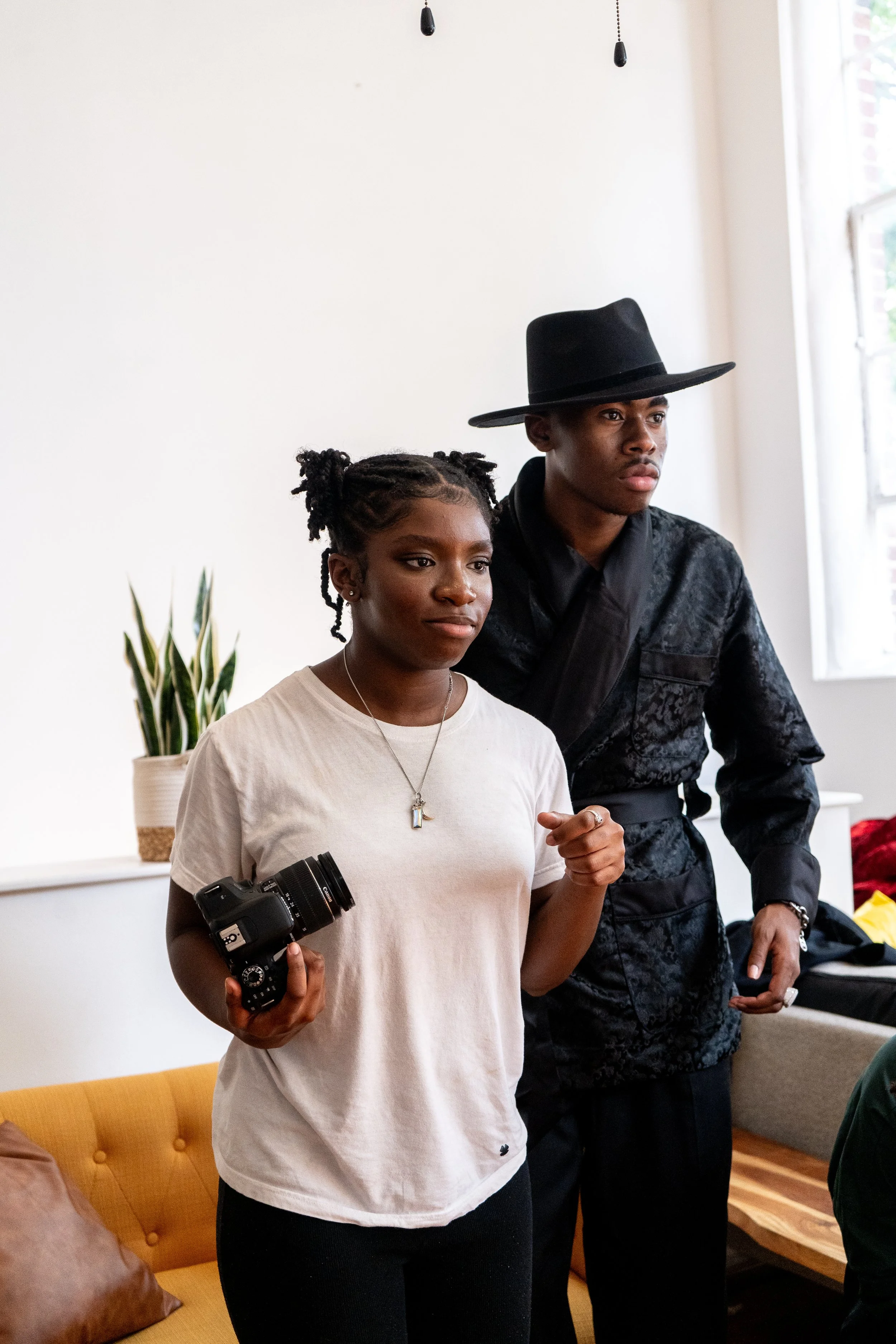 A woman with braids holding a camera and a man wearing a black hat standing next to her, indoors with a white wall, a plant, and a window in the background.