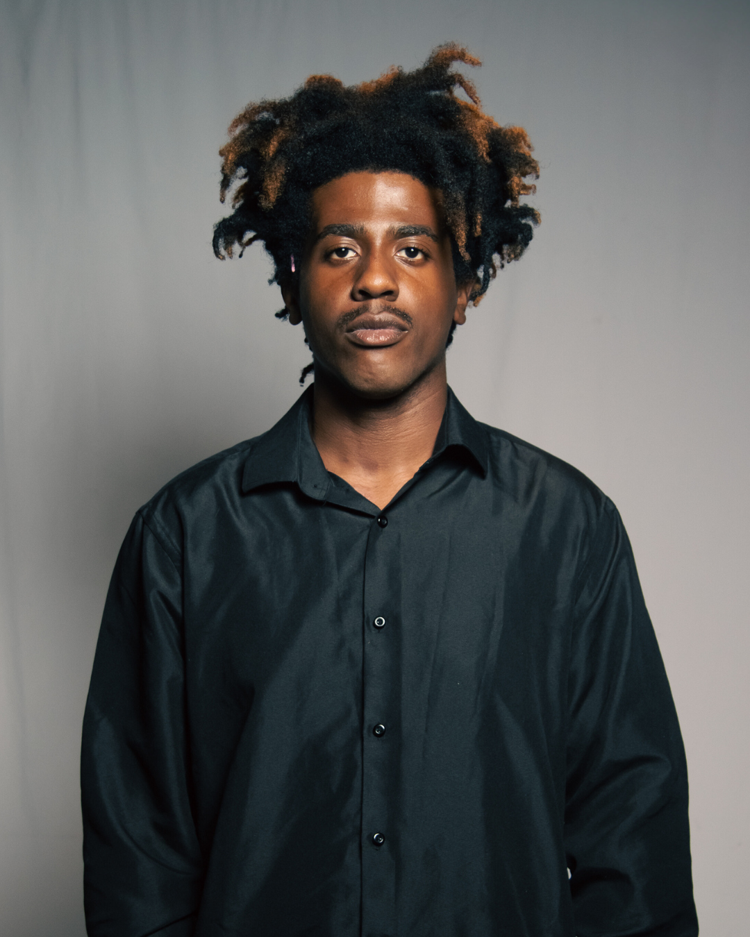 A young man with voluminous curly hair and a mustache wearing a black button-up shirt against a plain gray background.