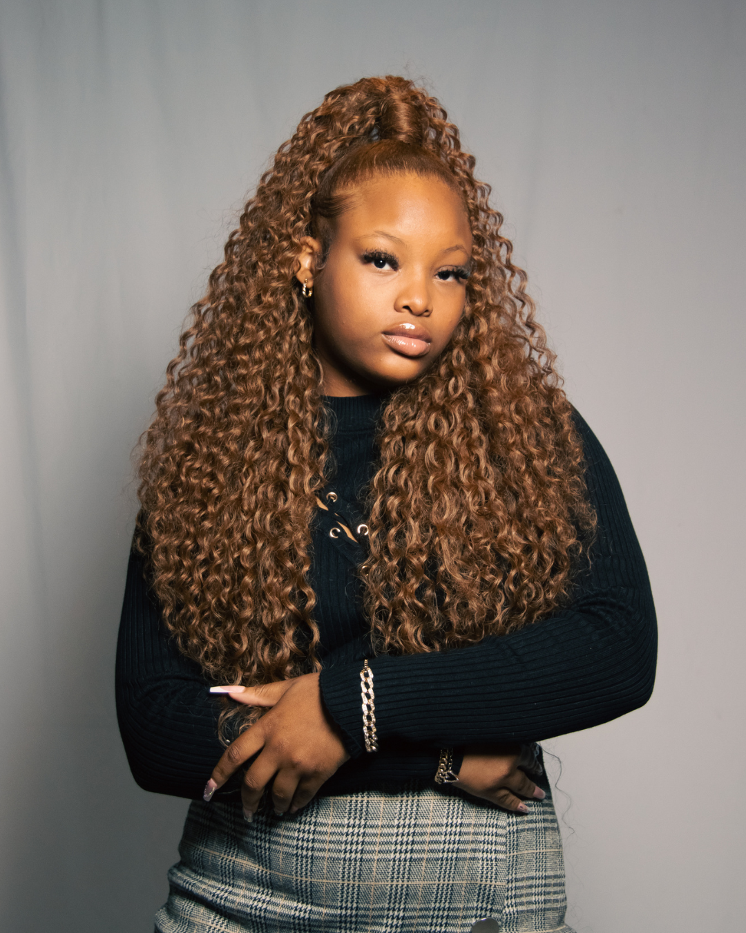 A woman with long, curly, light brown hair styled in a high ponytail, wearing a black ribbed sweater, plaid skirt, and jewelry, standing against a plain background.
