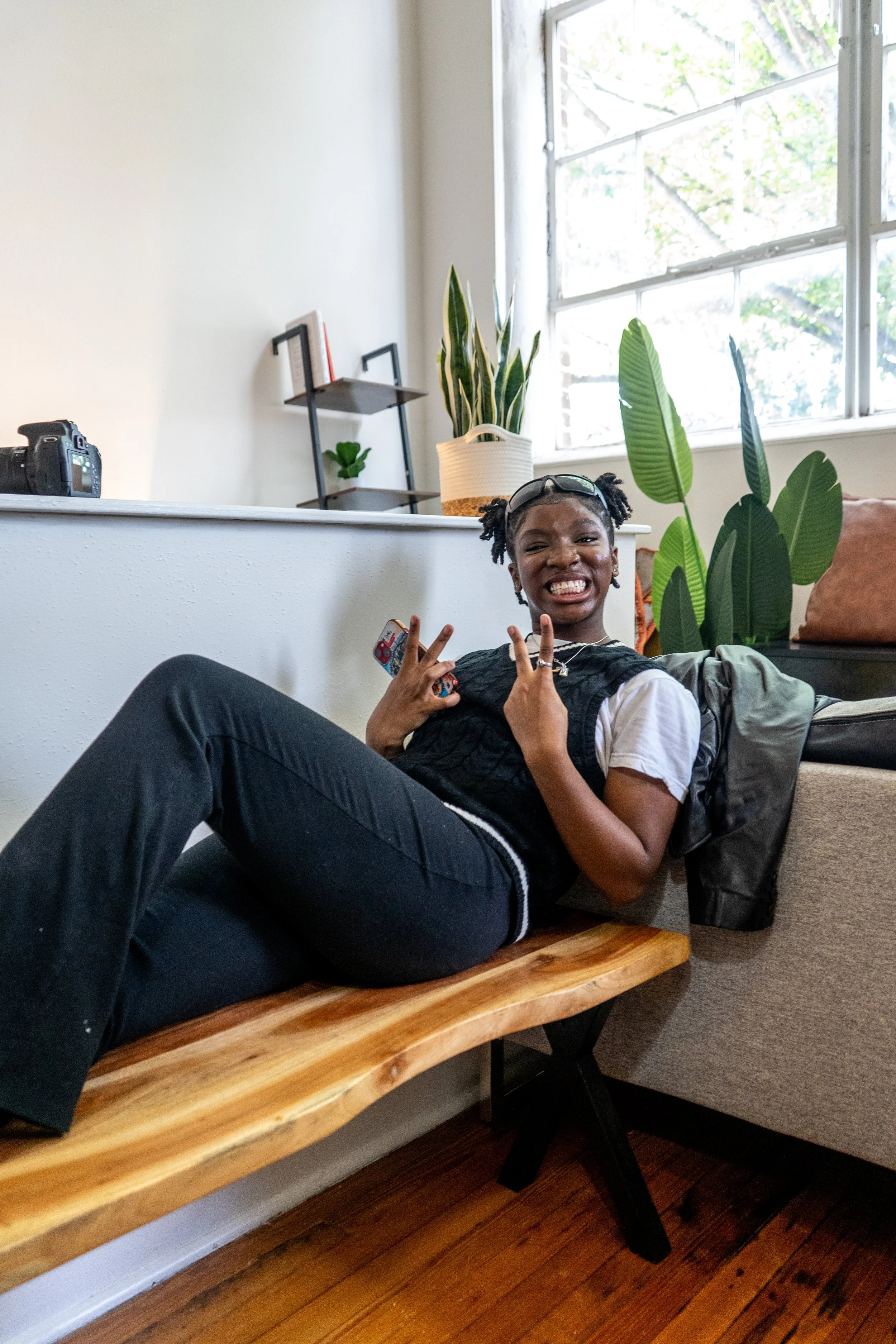 A girl with pigtails sitting on a wooden bench, smiling and making peace signs with her fingers inside a living room with plants and a window.
