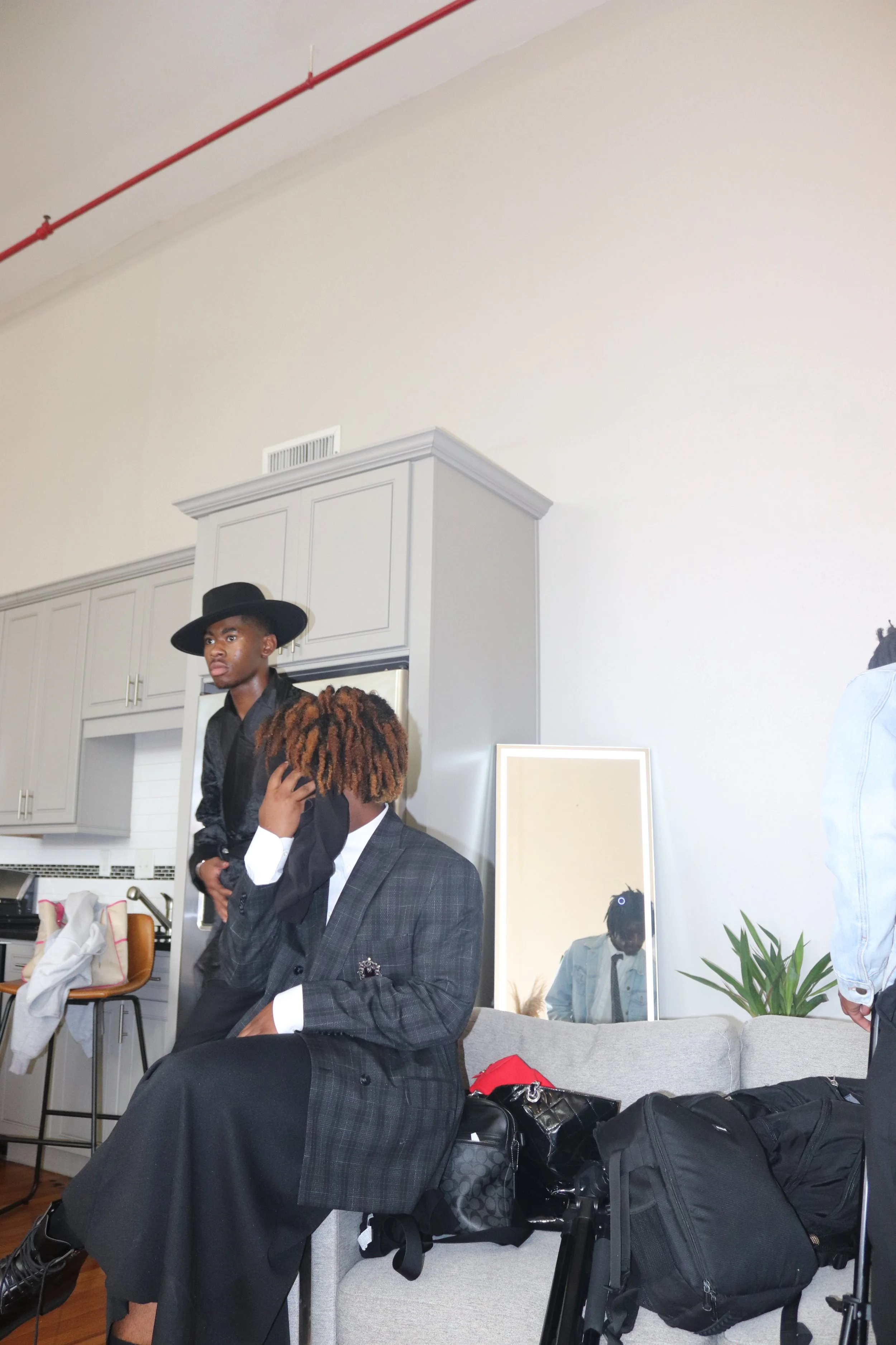 Two young men in fashionable clothing inside a modern living room, with one sitting on a sofa and talking on the phone, and the other standing nearby wearing a black hat. There are bags and a mirror behind them, a plant on the right side, and a mirro