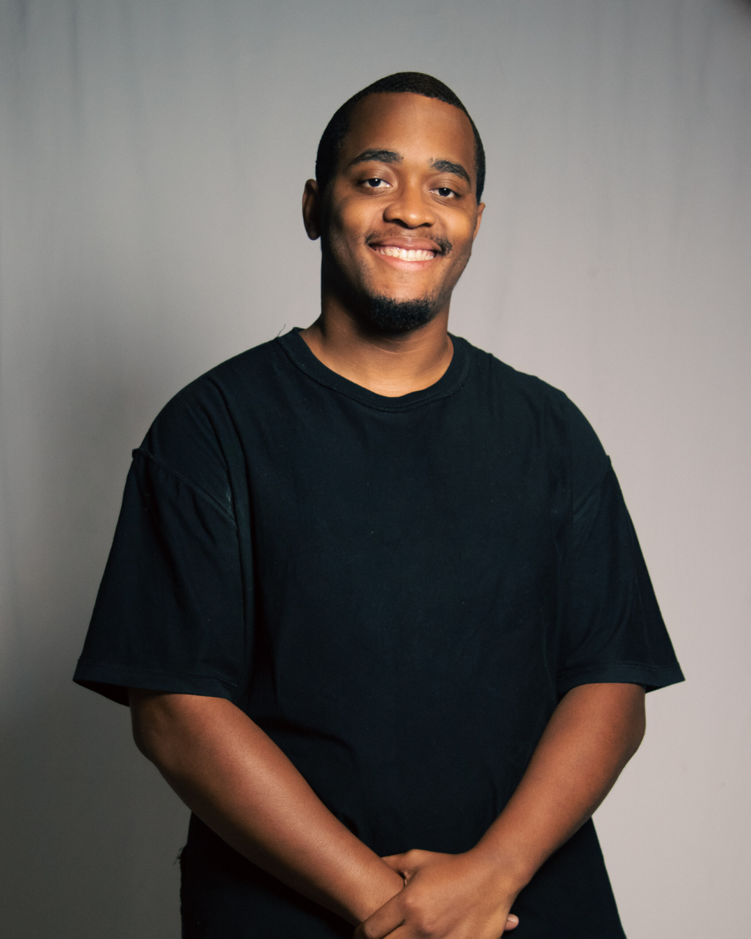 A young man with a short beard and short hair, smiling and wearing a black t-shirt, standing against a plain gray background.