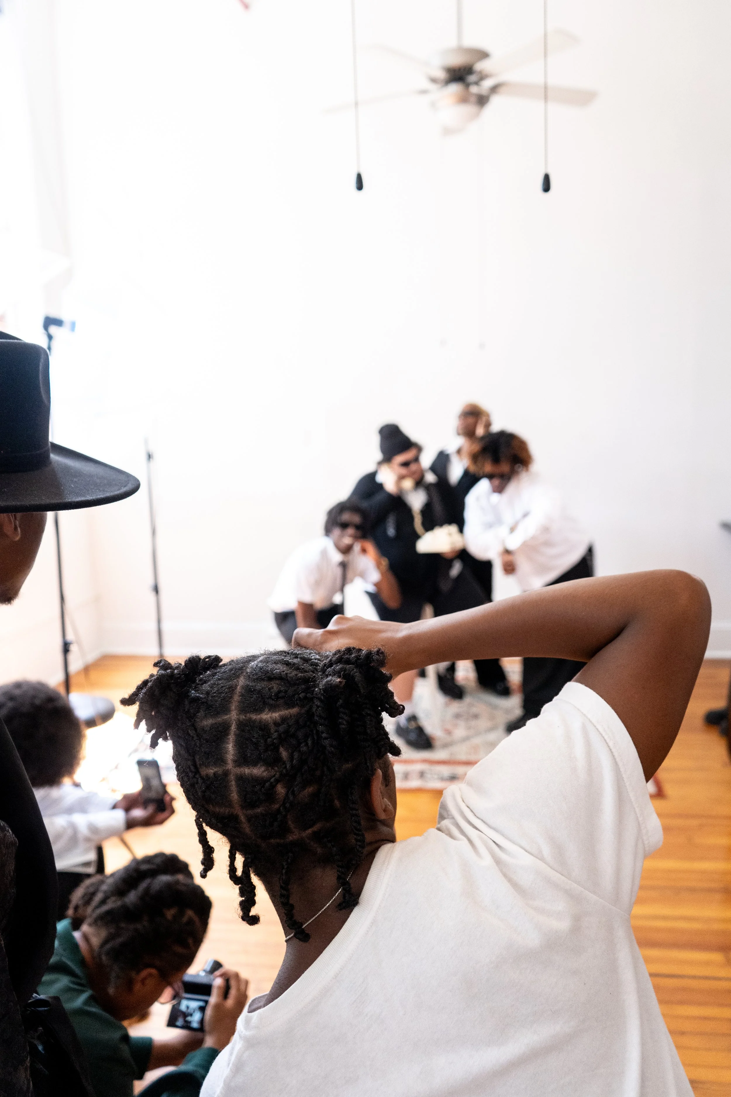 Group of people taking photos and filming a small photo shoot or scene inside a bright room with white walls, a ceiling fan, and hardwood floors.