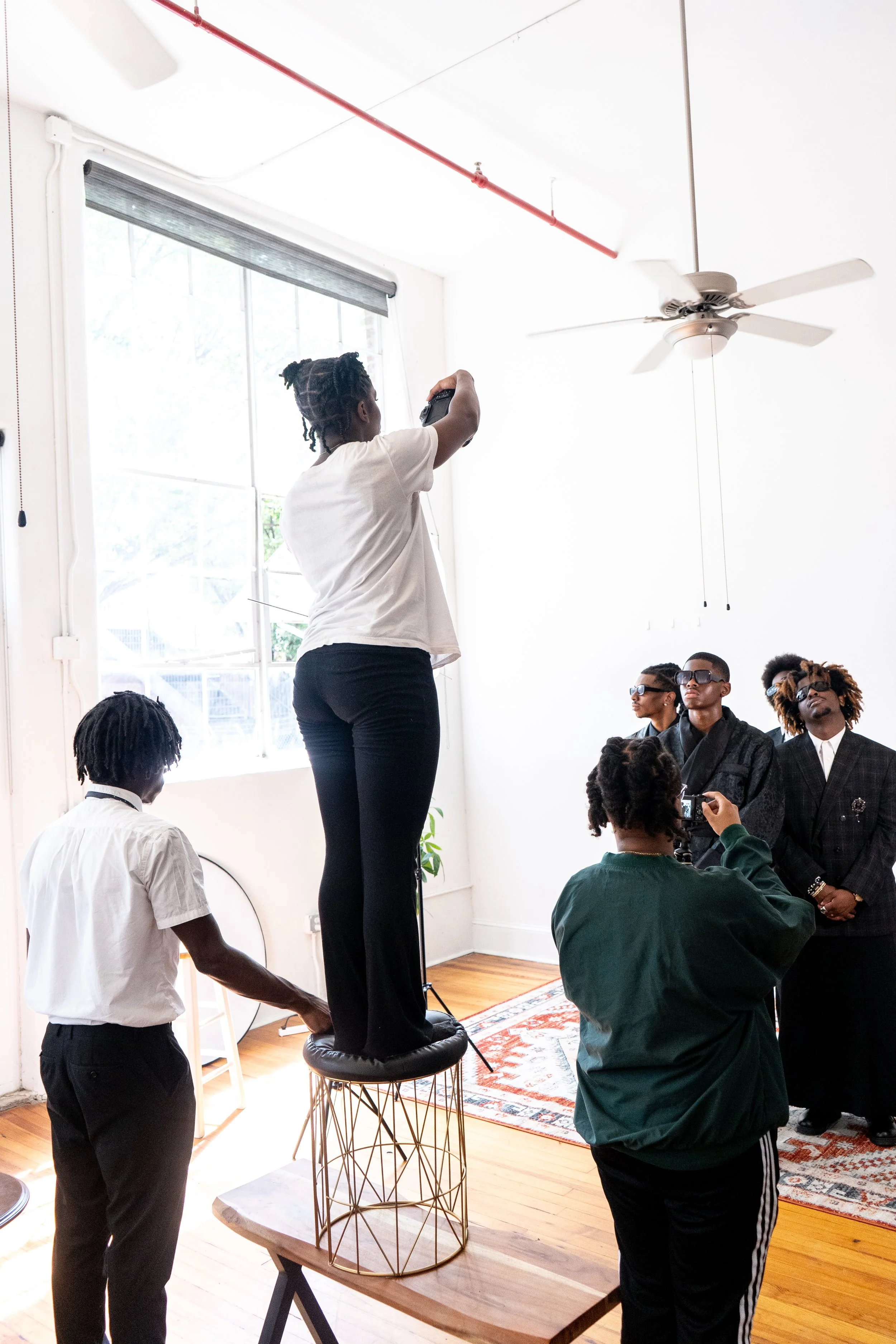 A woman standing on a round stool taking a photo of three men dressed in stylish black suits with sunglasses, while another person holds a camera and captures the scene in a brightly lit room with white walls and a large window.