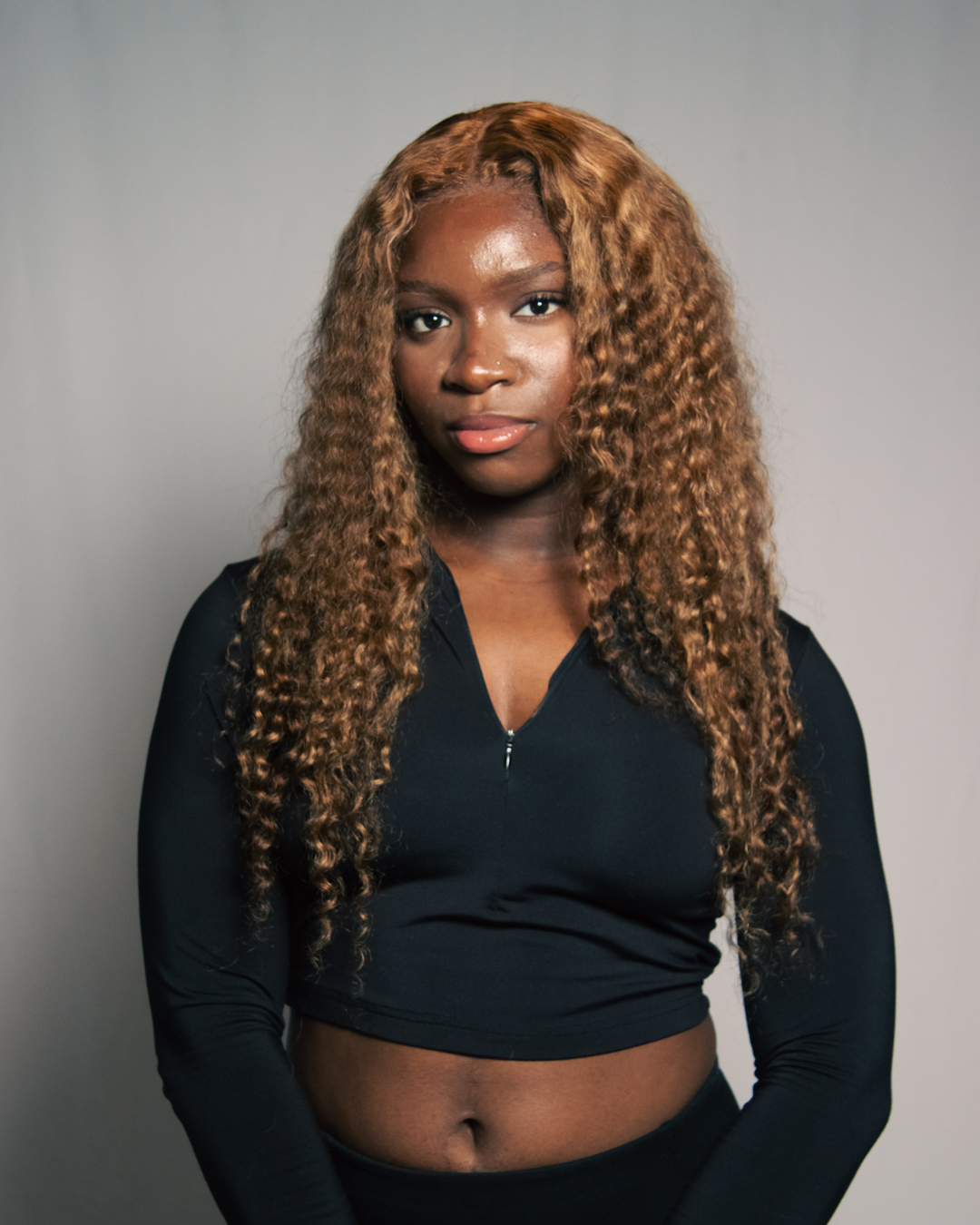A young woman with long, curly, light brown hair, wearing a black zip-up top and black bottoms, standing against a plain gray background.