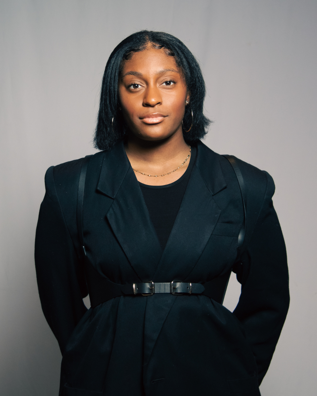 Portrait of a young African American woman with black hair, wearing a black blazer, a black top, hoop earrings, and a delicate necklace, standing against a neutral gray background.