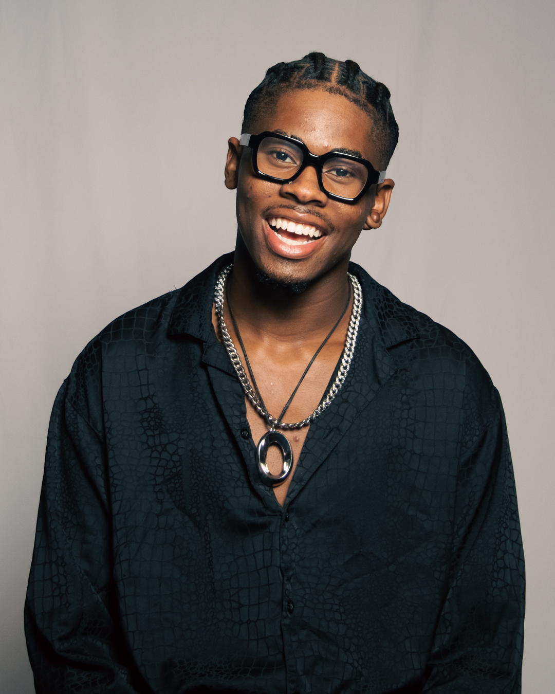 Portrait of a young man with glasses, dreadlocks, and a cheerful smile, dressed in a black shirt with a textured pattern, wearing layered necklaces, against a plain background.