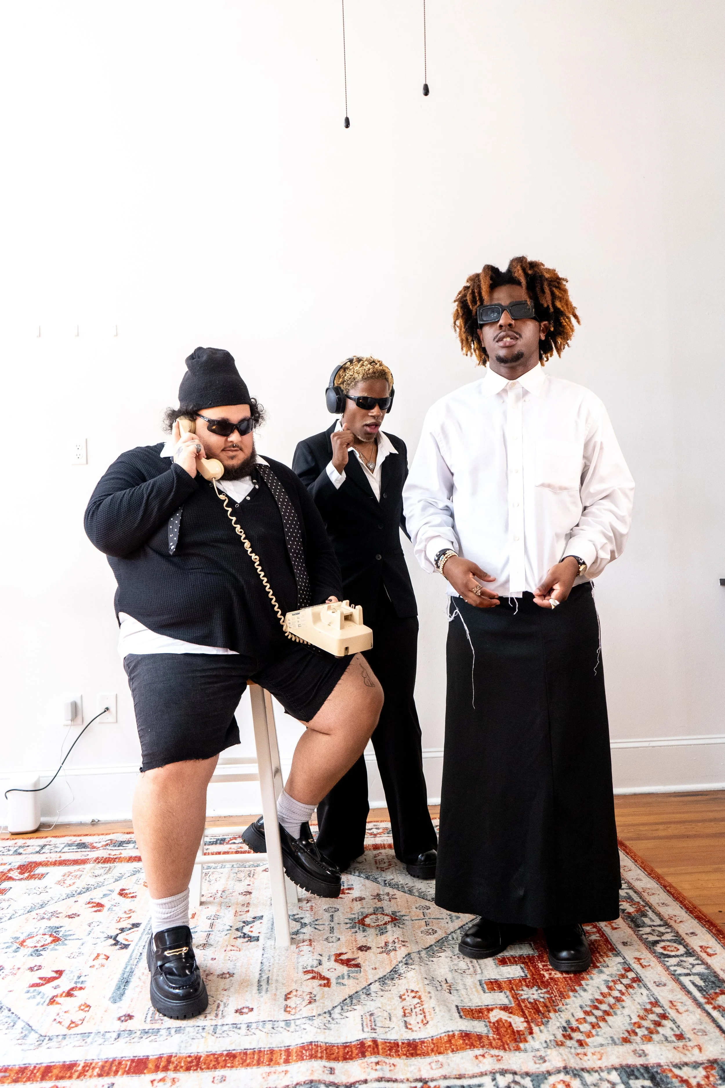 Three people with sunglasses in a room, one sitting on a stool with a vintage telephone, another speaking on a landline phone, and the third standing with hands in pockets.