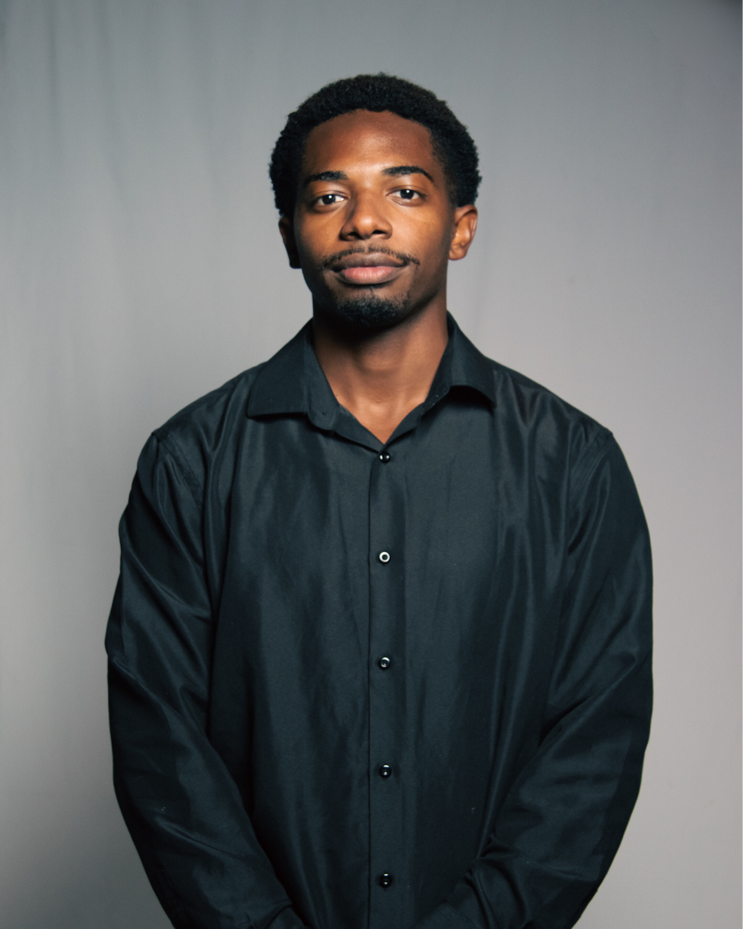 A young African American man with short curly hair, wearing a black button-up shirt, standing against a gray background, looking directly at the camera.