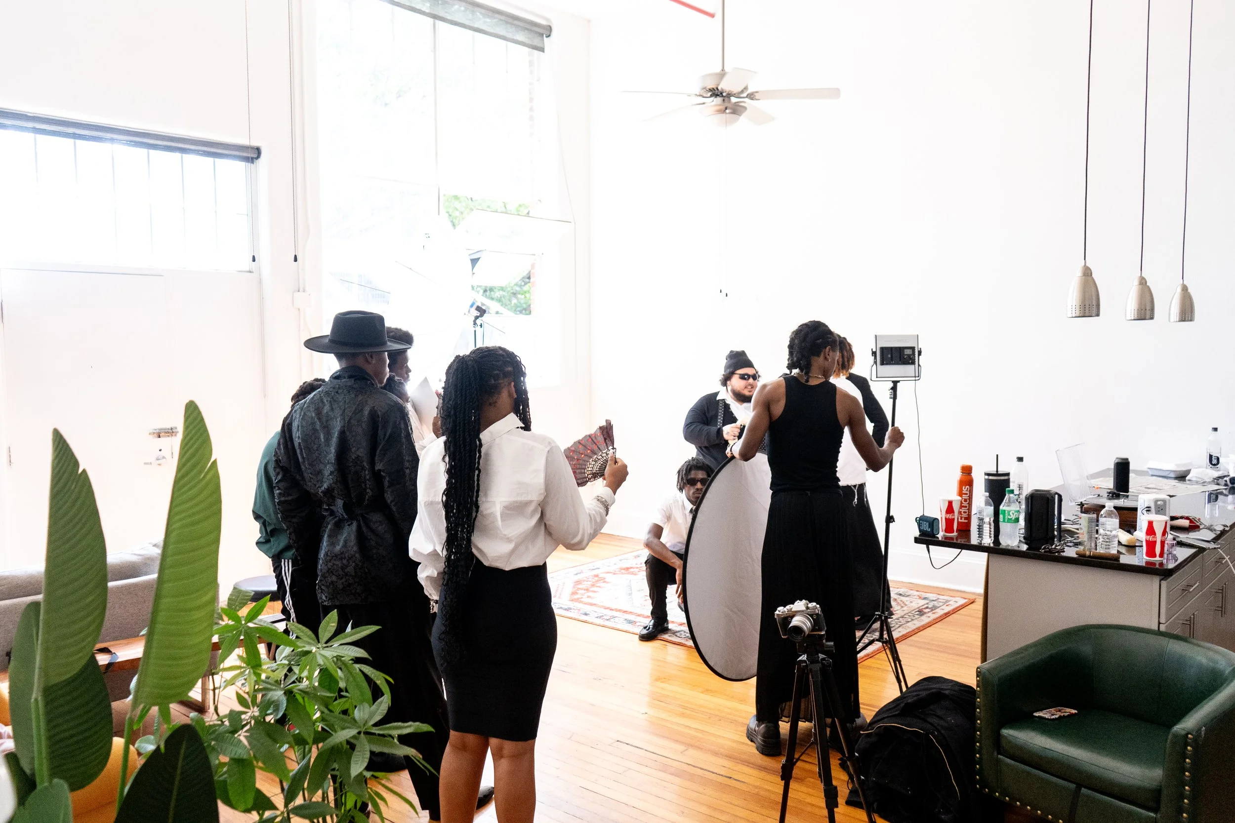 A group of photographers and crew working in a bright, modern indoor space with wooden floors, a green armchair, and large windows. They are preparing for a photoshoot with professional equipment, lighting, and various supplies on the counter.