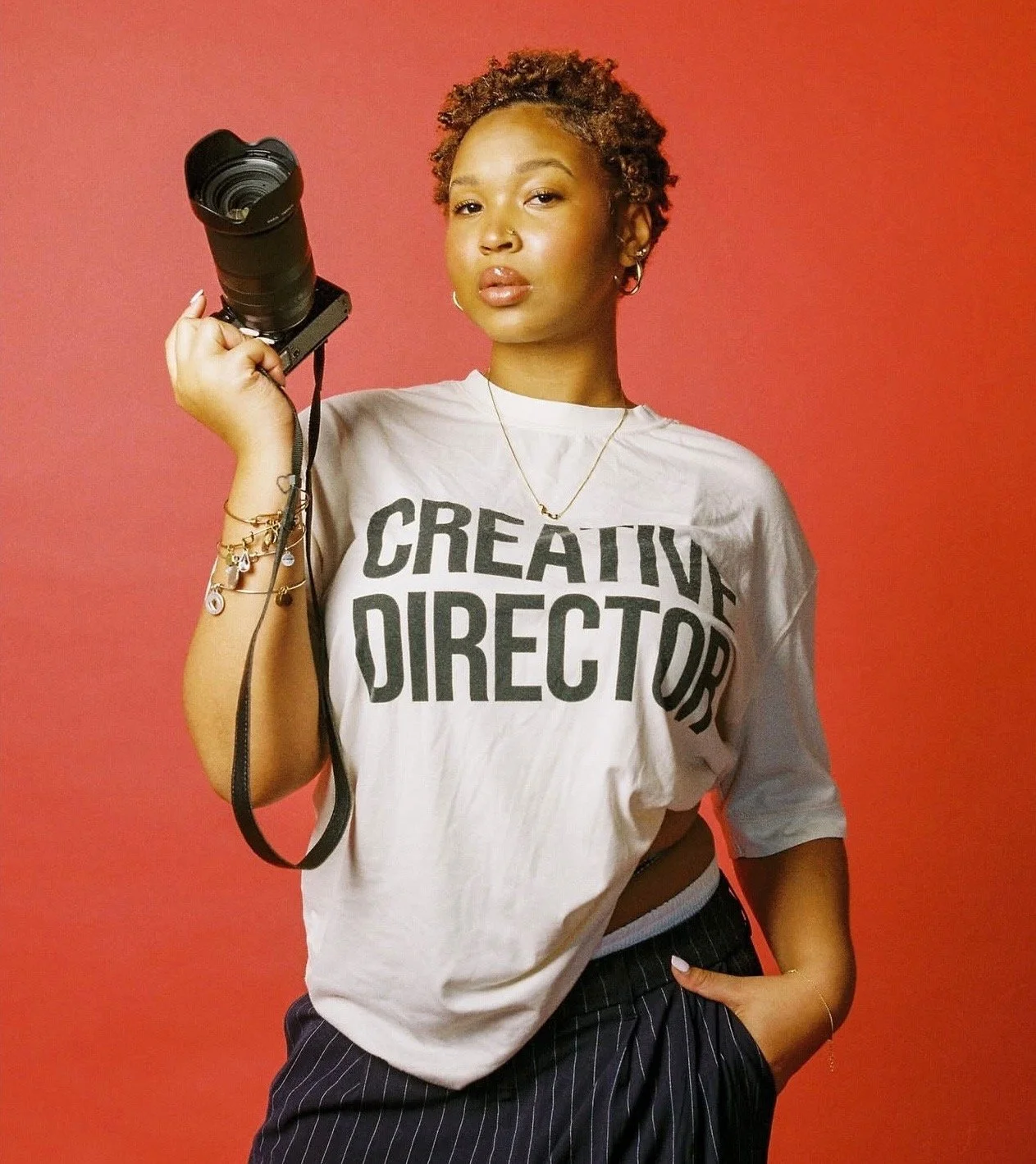 Young woman with short curly hair holding a camera, wearing a white T-shirt that says 'CREATIVE DIRECTOR', against a red background.