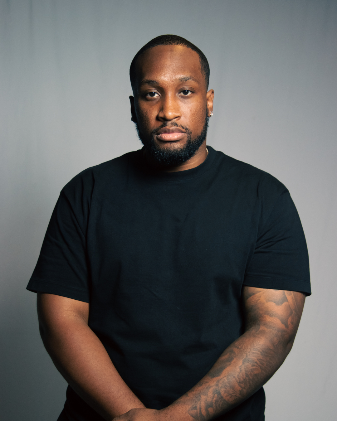 A young Black man with a close-cropped beard and mustache, wearing a black T-shirt, standing against a plain gray background, looking directly at the camera with a serious expression.