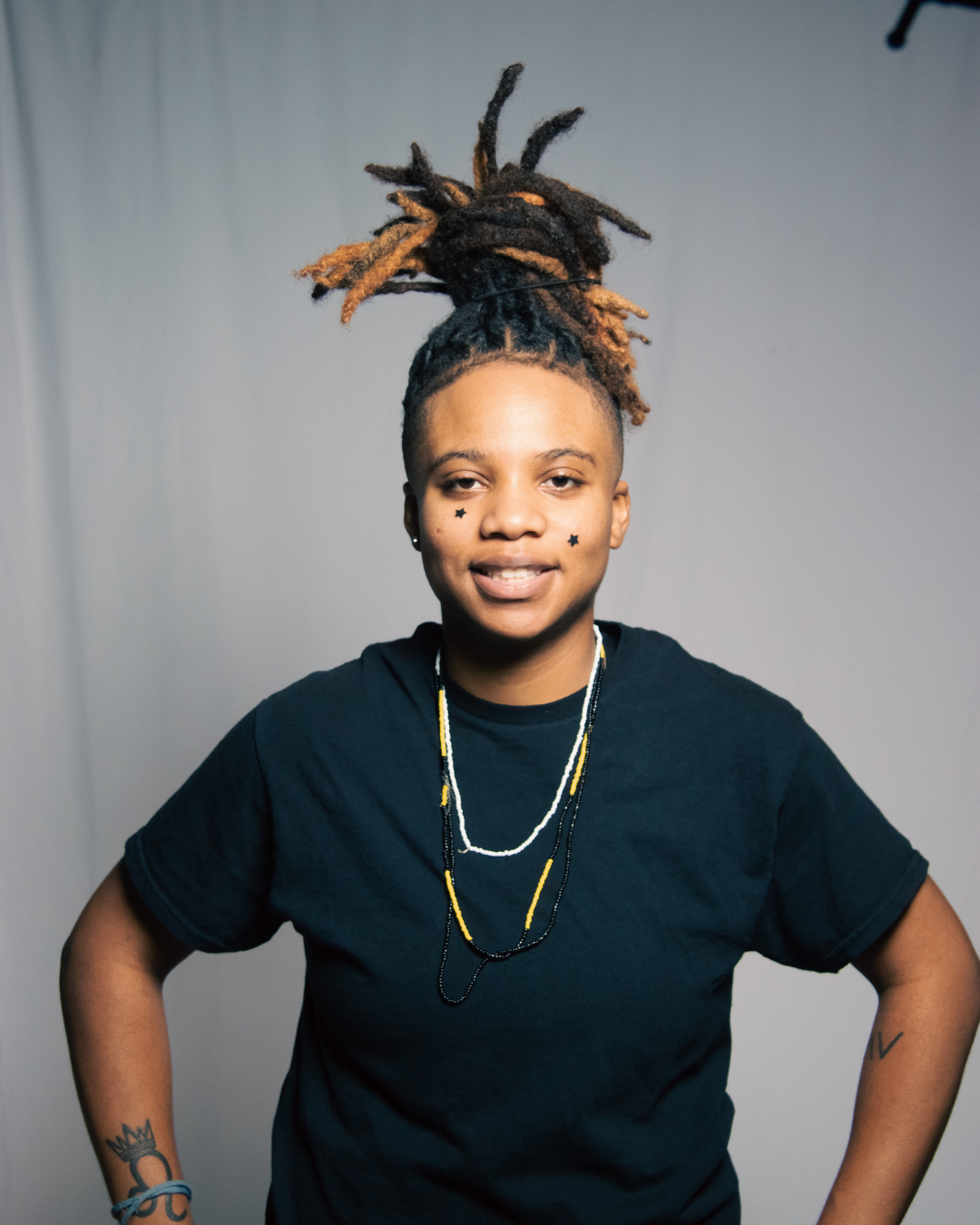A young person with dreadlocks styled in an updo, wearing black star face stickers, a black t-shirt, layered necklaces, and tattoos on their arms, standing in front of a plain background.