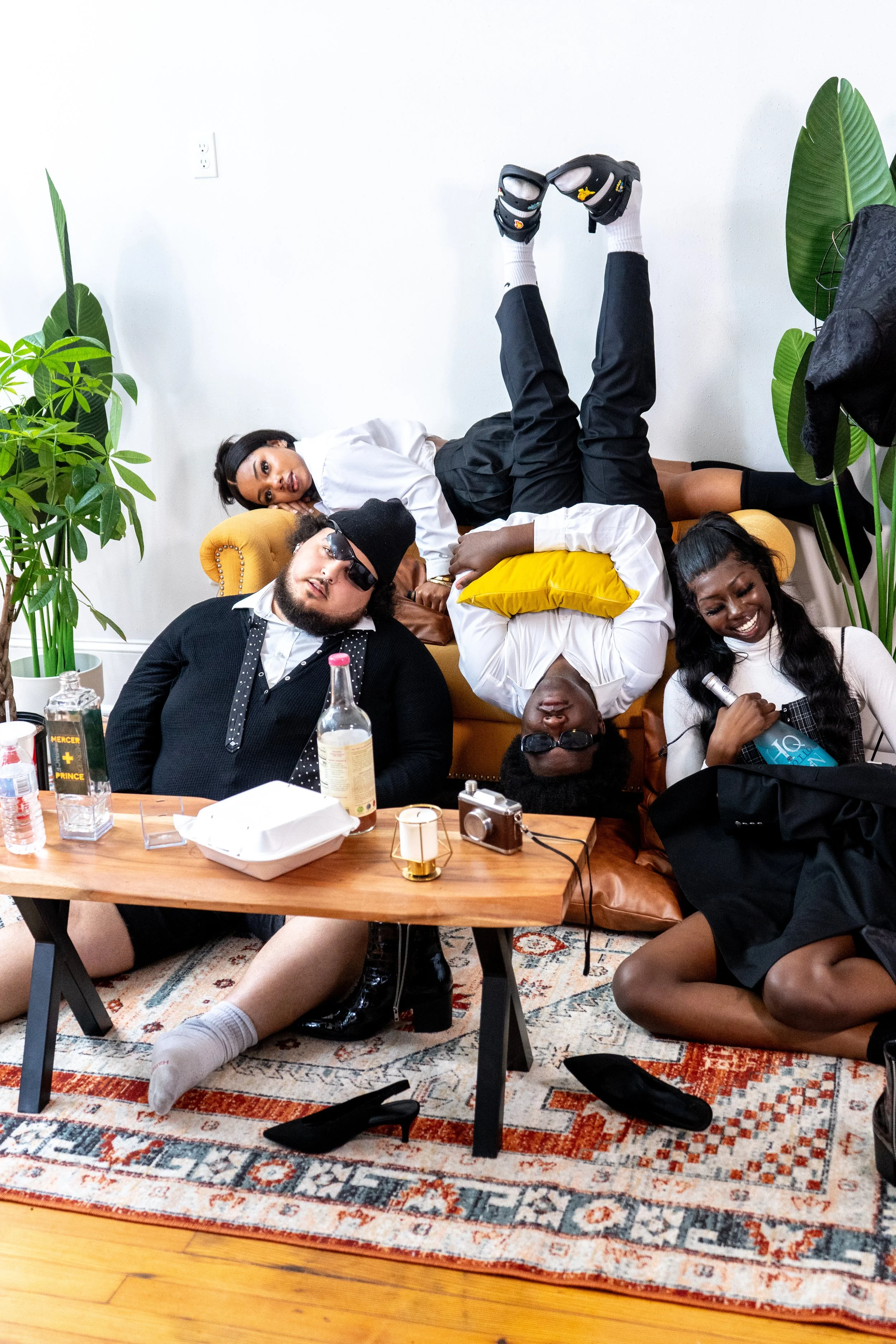 Four young people relaxing and posing indoors, with some sitting on and others lying on a yellow couch, surrounded by plants and a wooden coffee table with drinks, snacks, cameras, and a microphone.