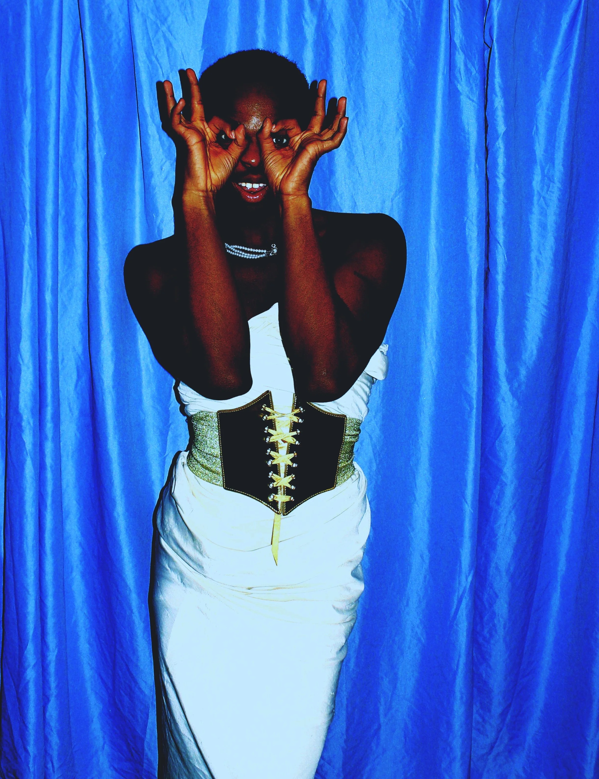 Person wearing a white dress with a black and gold corset, posing in front of a blue curtain, making a playful gesture with their hands around their eyes.