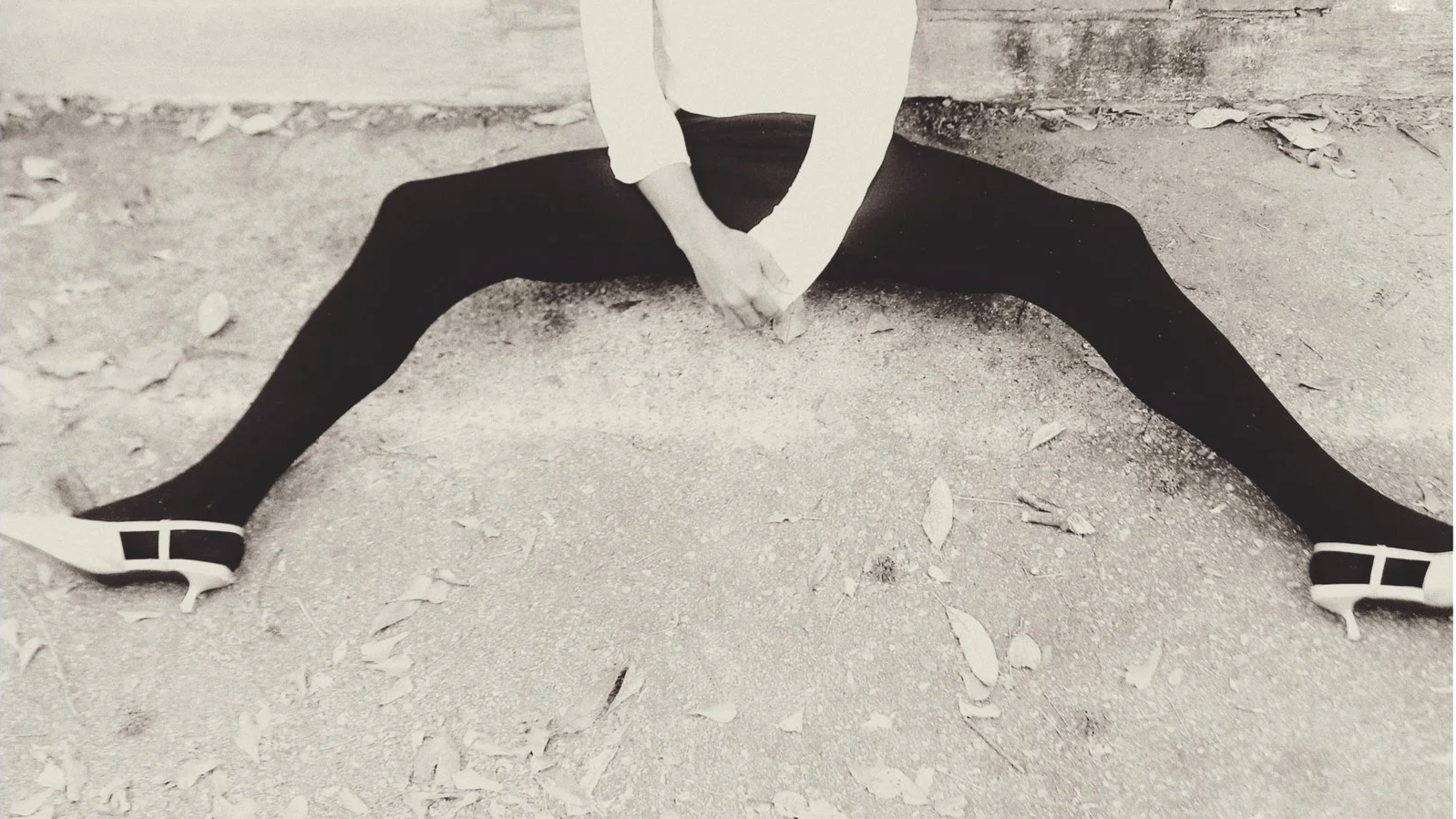 Person sitting on the ground with legs spread wide apart, wearing black leggings, white shoes, and a white top, with hands clasped between legs, outdoors on dirt with fallen leaves.