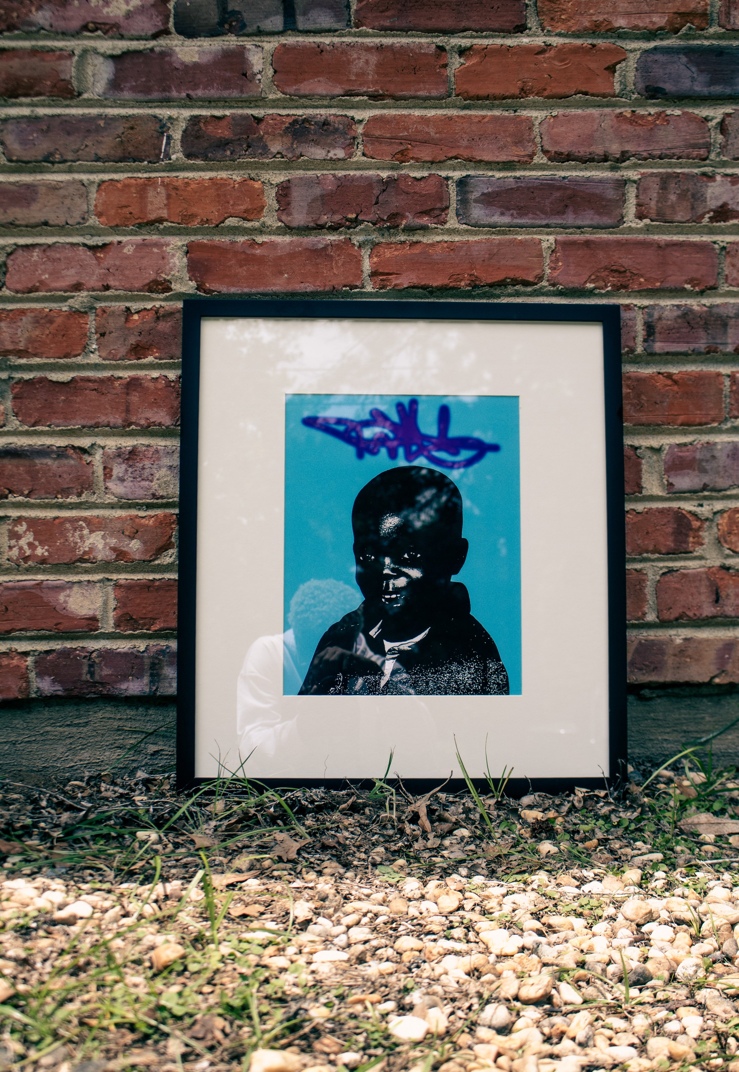 Framed black and white photograph of a person with glasses and a beanie, displayed outdoors against a brick wall.