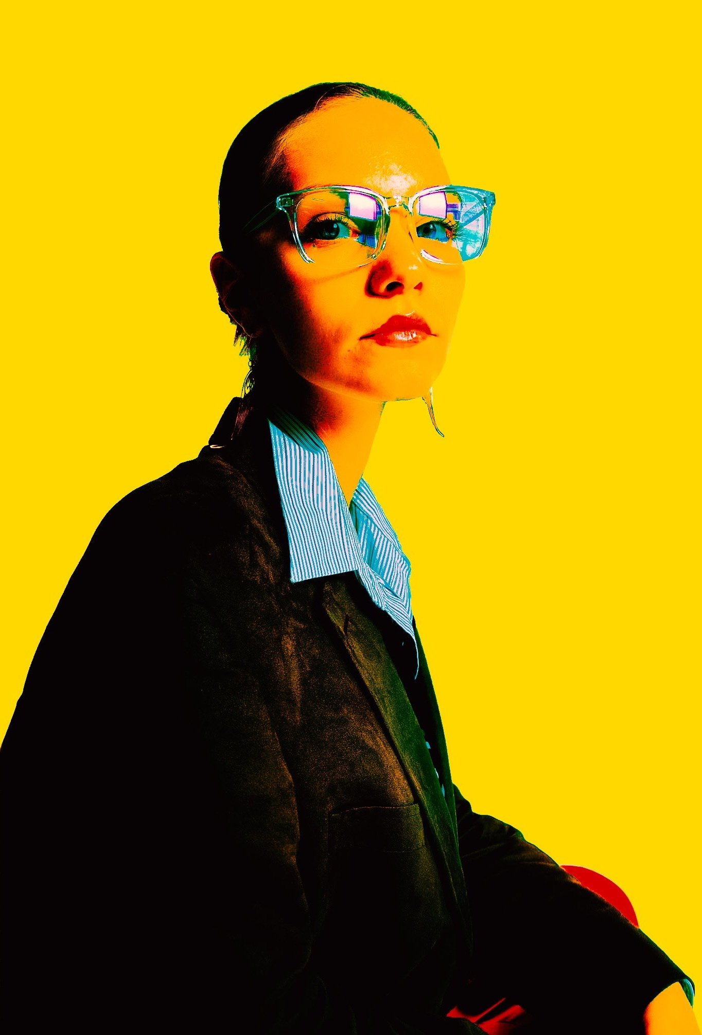 A woman with slicked-back hair and glasses with a reflective blue tint, wearing a striped shirt and black blazer, against a vibrant yellow background.