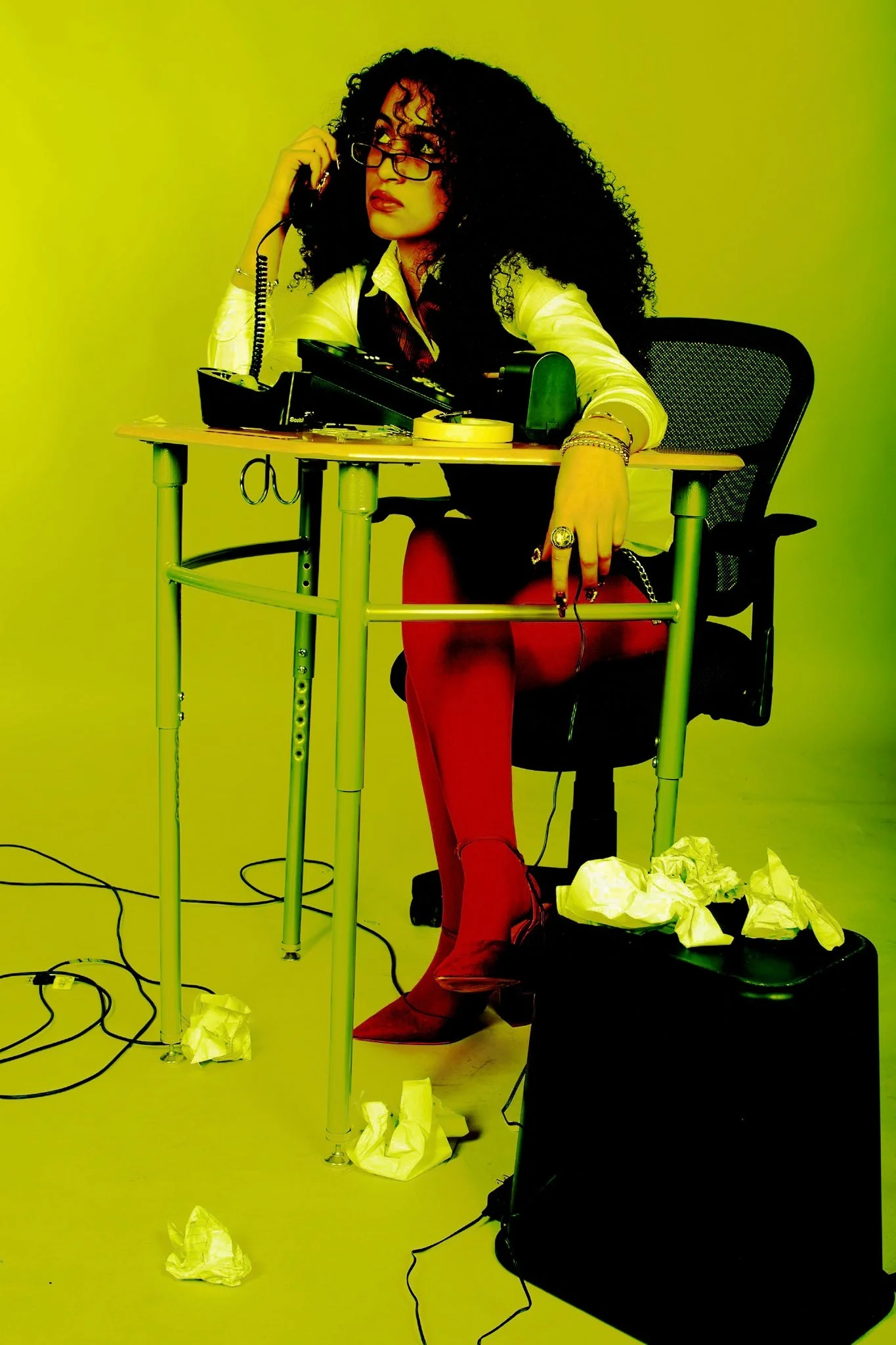 A woman with curly black hair, glasses, and red lipstick, sitting at a disorganized desk with crumpled papers and tangled cords. She is wearing a white shirt, red tights, and pointed shoes, and appears to be on a phone call.