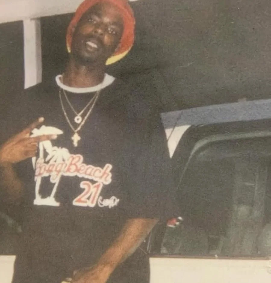 A man wearing a colorful hat, glasses, and layered necklaces, pointing to his shirt that says 'Beach 21' and making a peace sign with his hand.