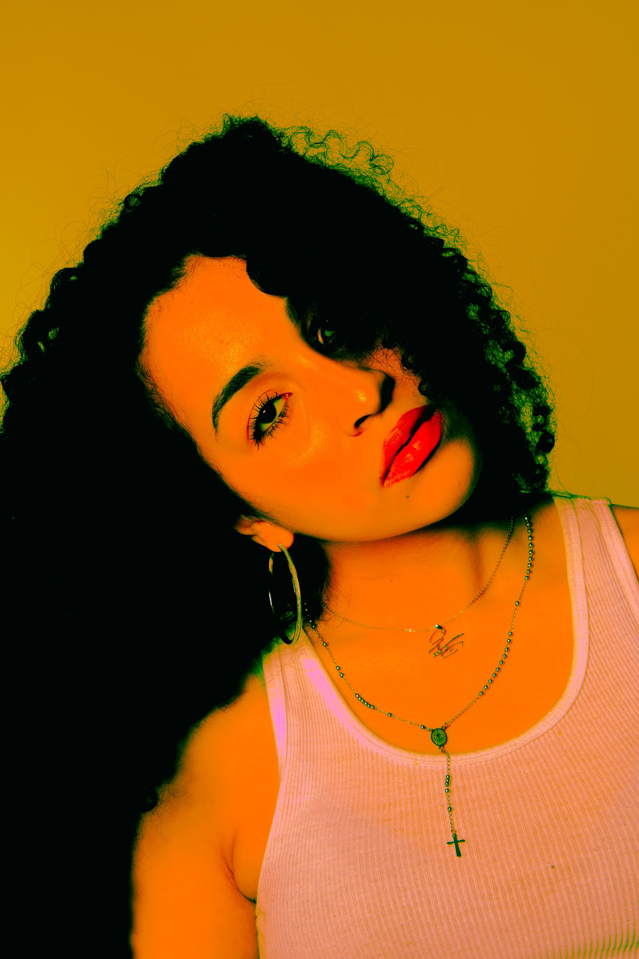 A woman with curly dark hair wearing a light pink tank top, hoop earrings, and layered necklaces including a cross pendant, posing against a yellow background.