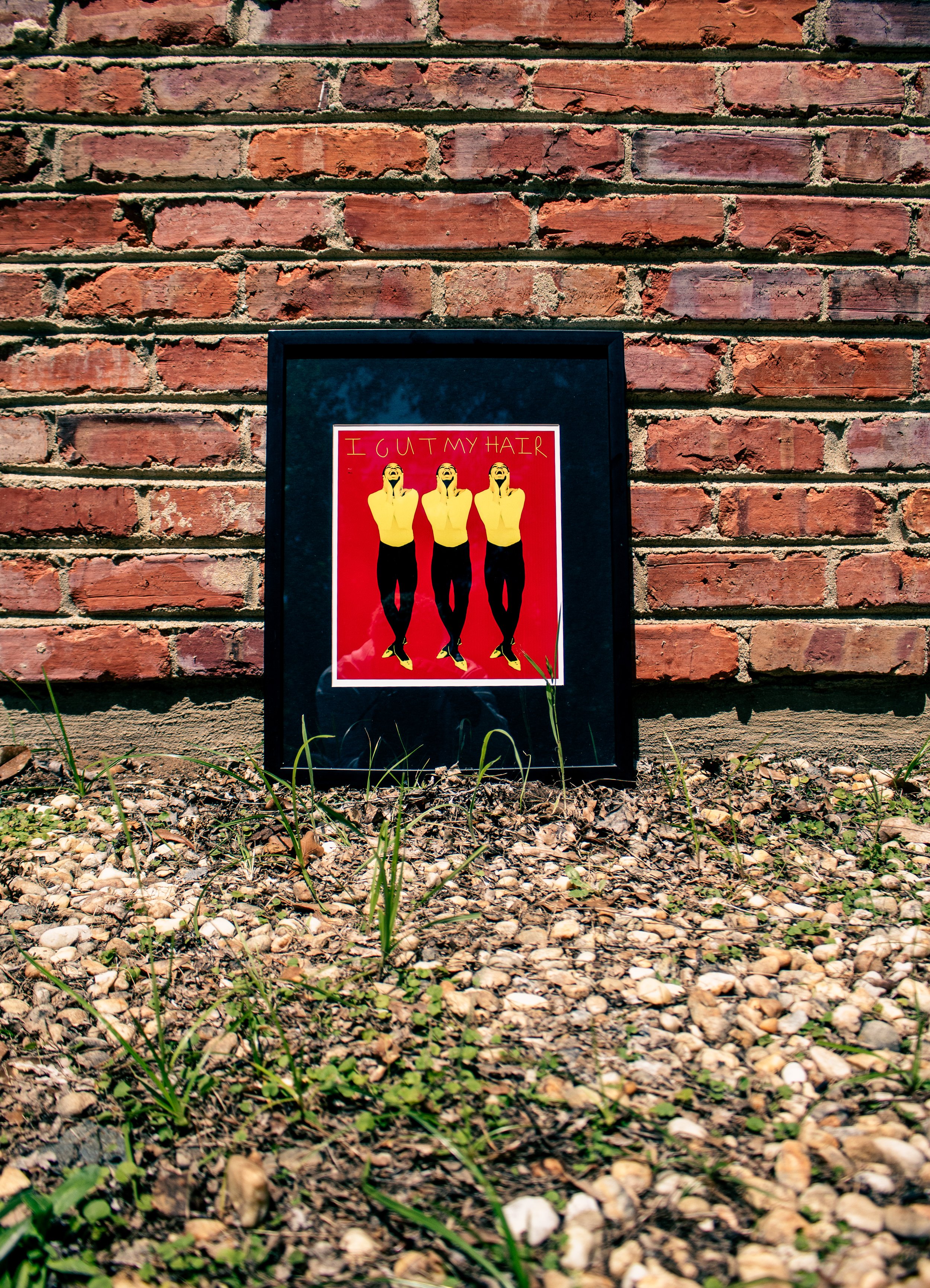 Framed poster leaning against a brick wall, showing three identical women in yellow tops and black pants, with the text "I GUT MY HAIR" at the top, on a background of red.