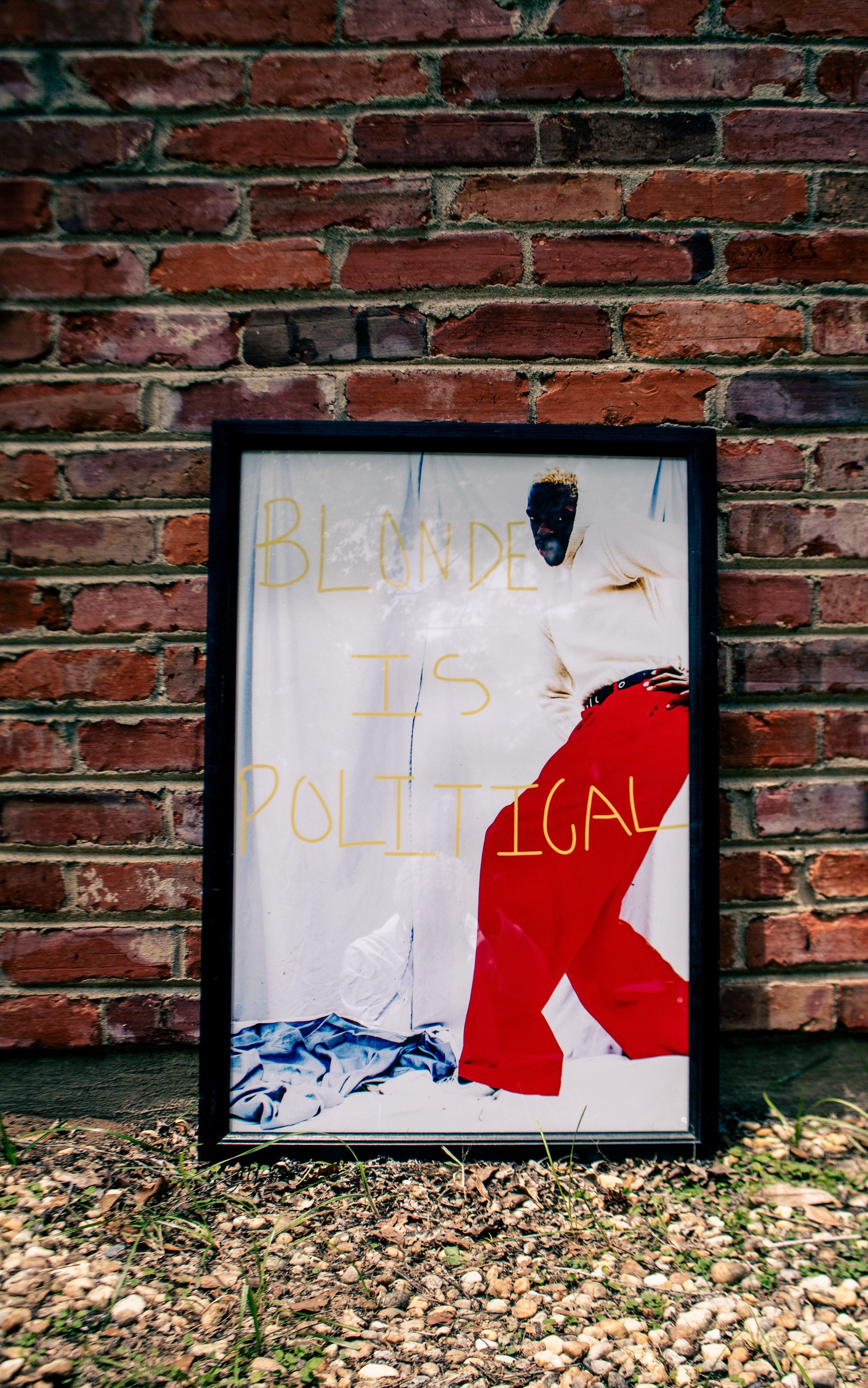 A framed poster leaning against a brick wall, depicting a person with dark skin and short blonde hair, wearing a white shirt and red pants, with the words "BLONDE IS POLITICAL" written in yellow across the poster.