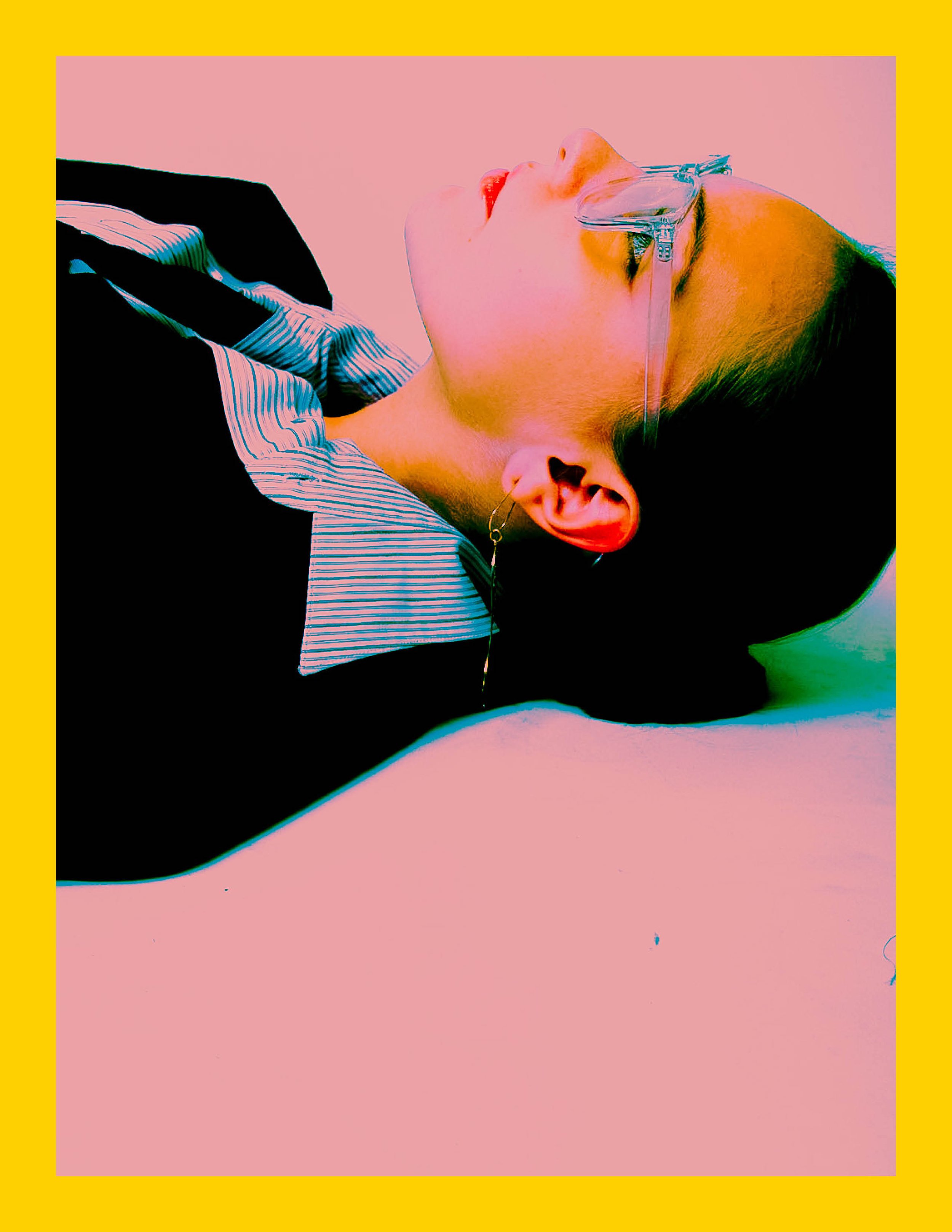 Person lying on pink surface with glasses and striped shirt, with a drop of water on their face.
