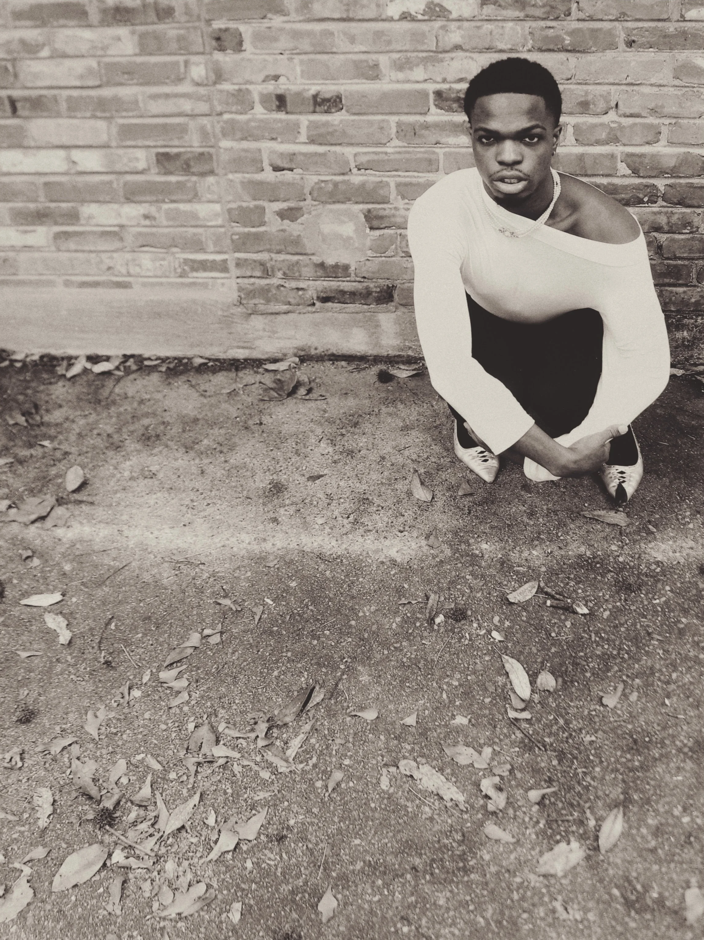 A young man squatting on the ground in front of a brick wall, wearing a off-the-shoulder top, dark pants, and sneakers, looking directly at the camera.