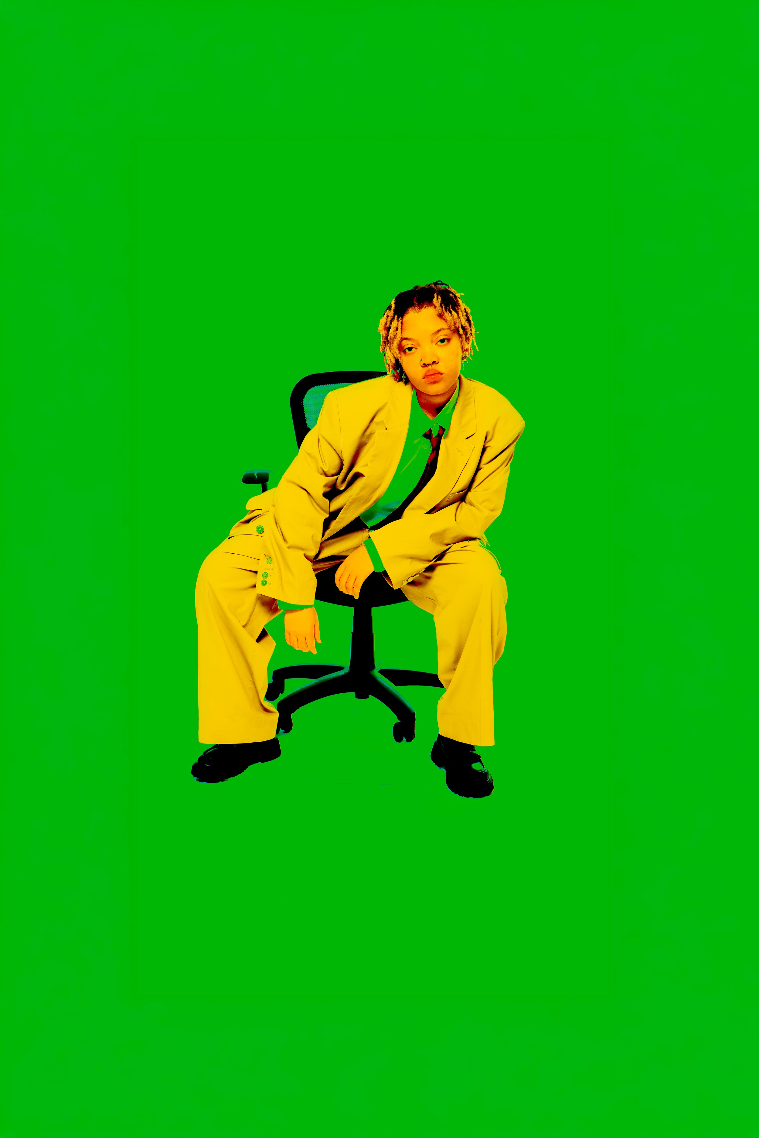 A person with short dreadlocks, wearing a yellow suit and green shirt, sitting on a black office chair against a solid green background.