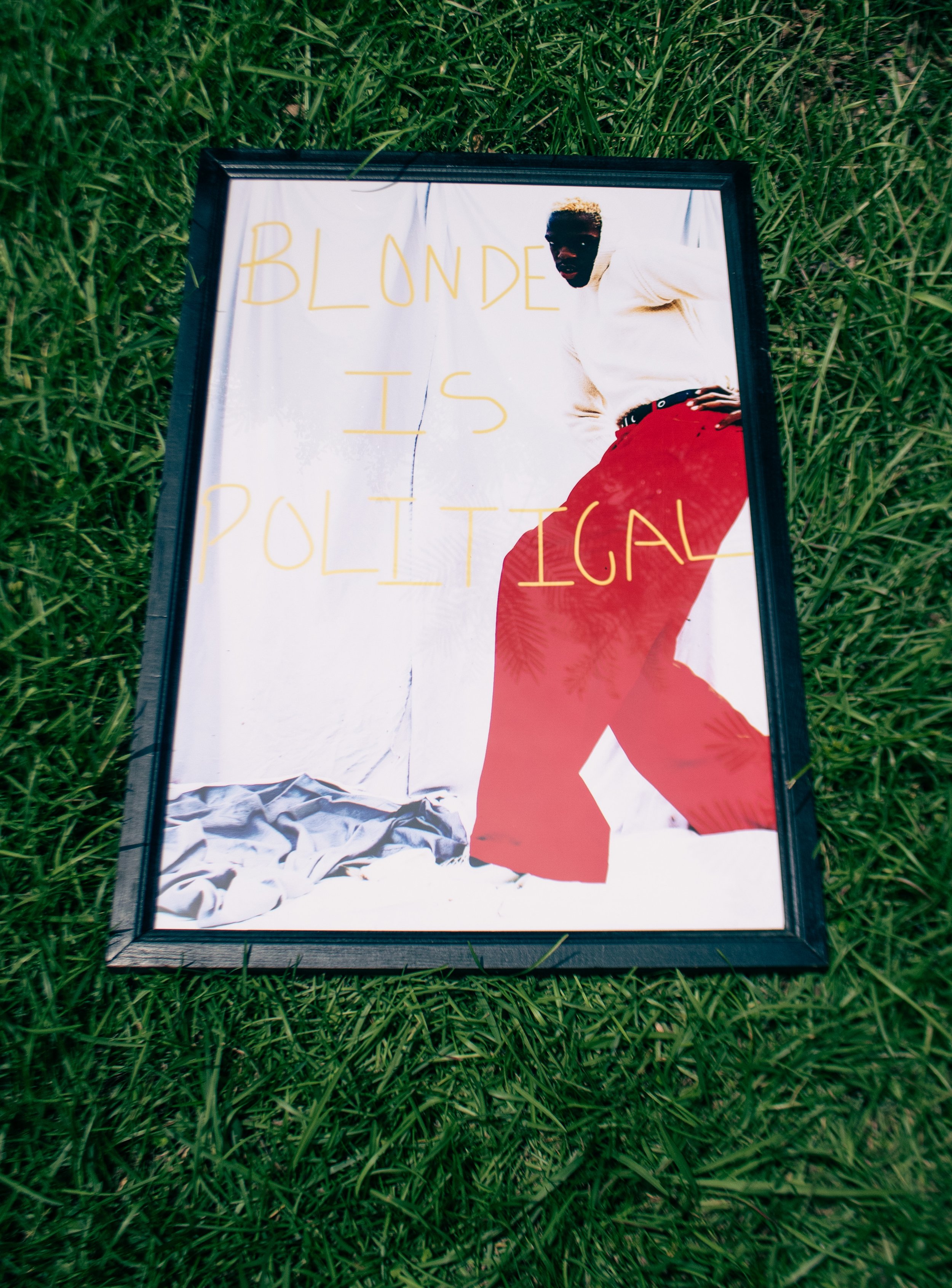 Framed poster on grass featuring a woman in a white top and red pants, with the words 'Blonde is Political' written in yellow.