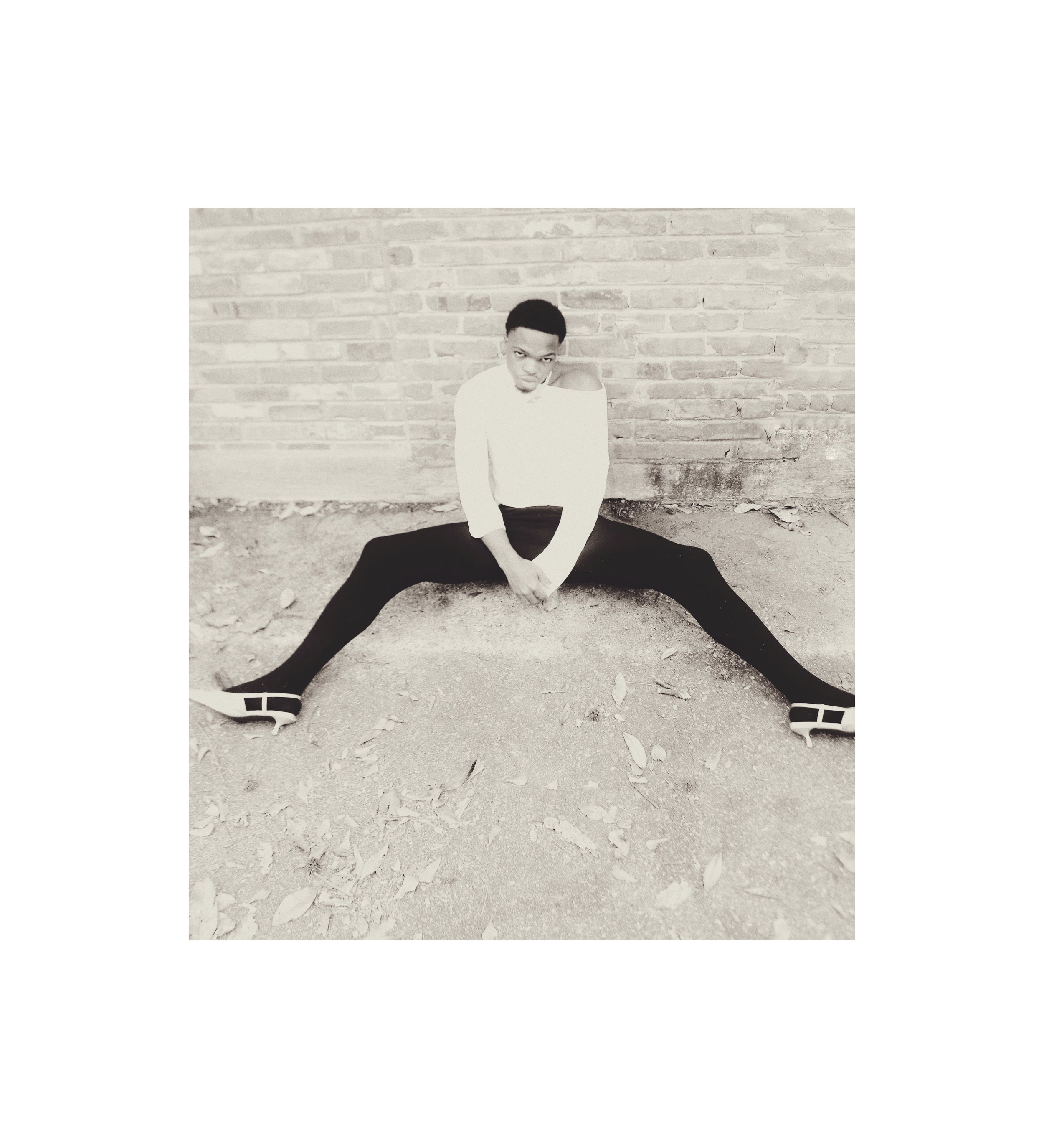 Young man sitting on the ground in front of a brick wall, legs spread wide apart, wearing a white shirt, black pants, and shoes, with a serious expression.