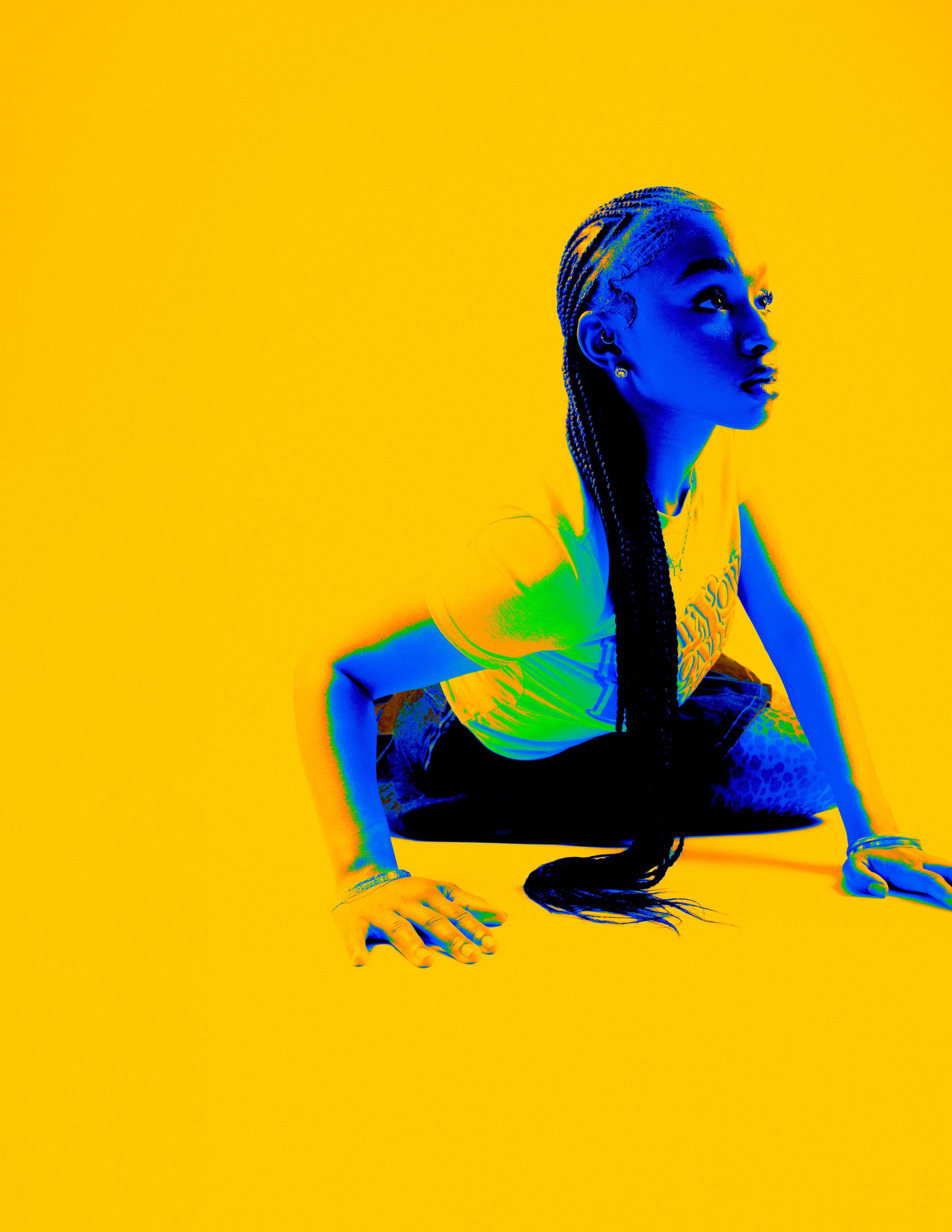 A woman with long braided hair crouches on the floor against a yellow background, with a colorized, high-contrast effect.