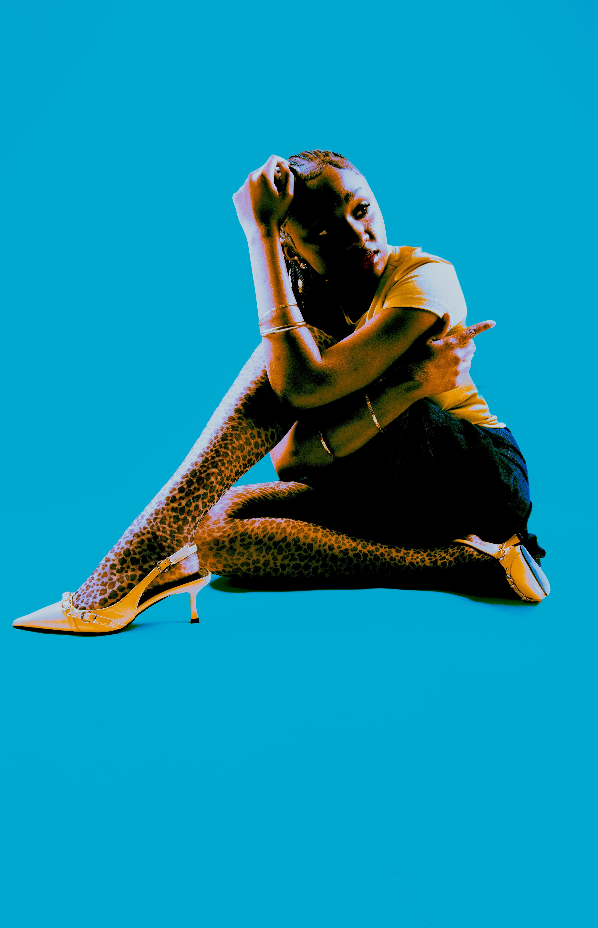 A woman with braided hair, wearing a yellow top and leopard print tights, sits on the floor against a solid blue background. She wears yellow high heels and poses with her hand on her forehead and her other arm across her chest.