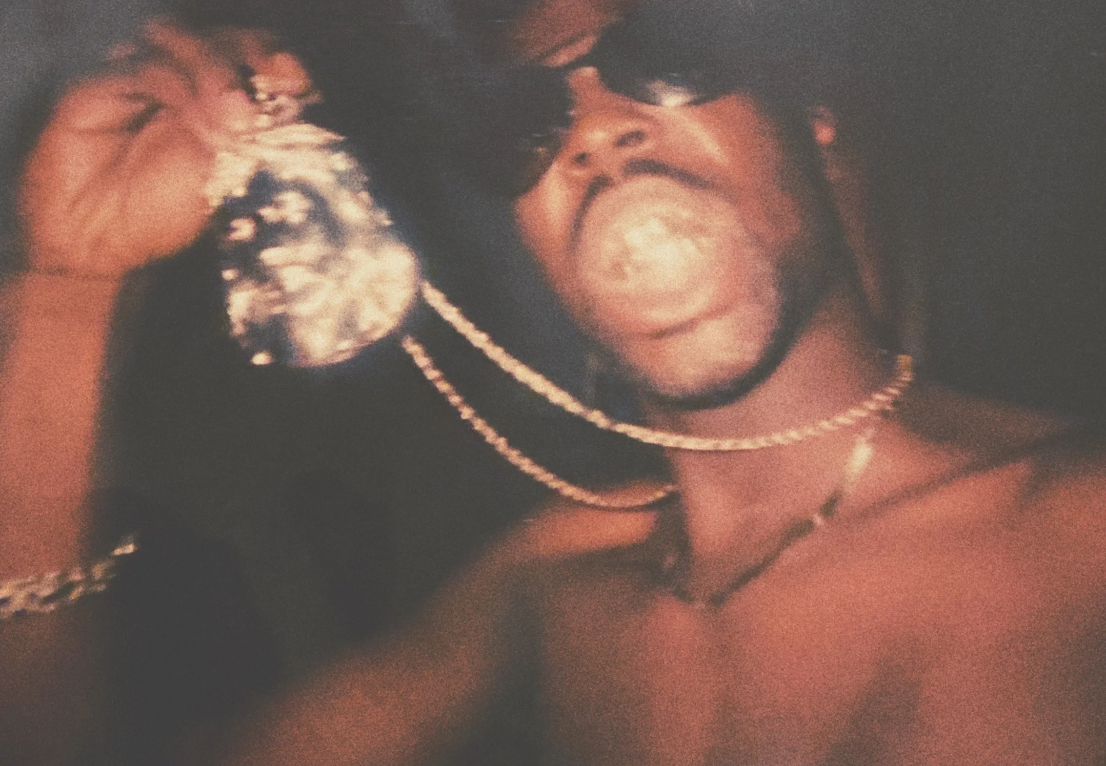 A young man with dreadlocks and wearing sunglasses, silver jewelry including a chain, a bracelet, and a necklace, holding a large diamond ring close to his face and making a playful expression.