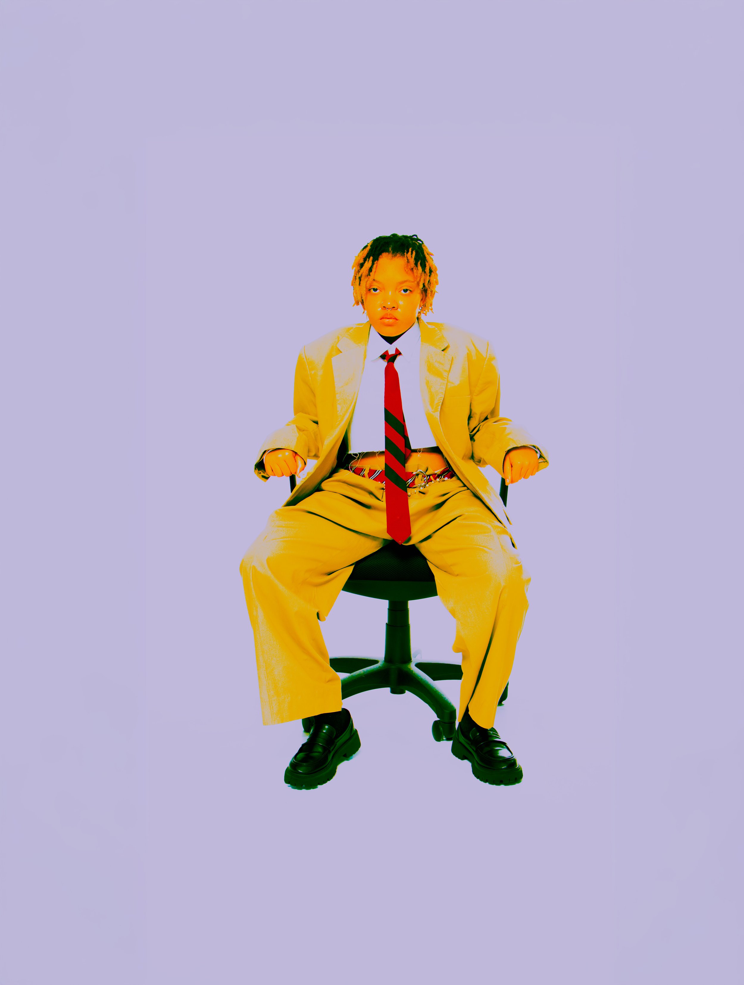 A young person with dreadlocks sitting on a black office chair, wearing a yellow suit, white shirt, red and black striped tie, and black shoes, against a plain light purple background.