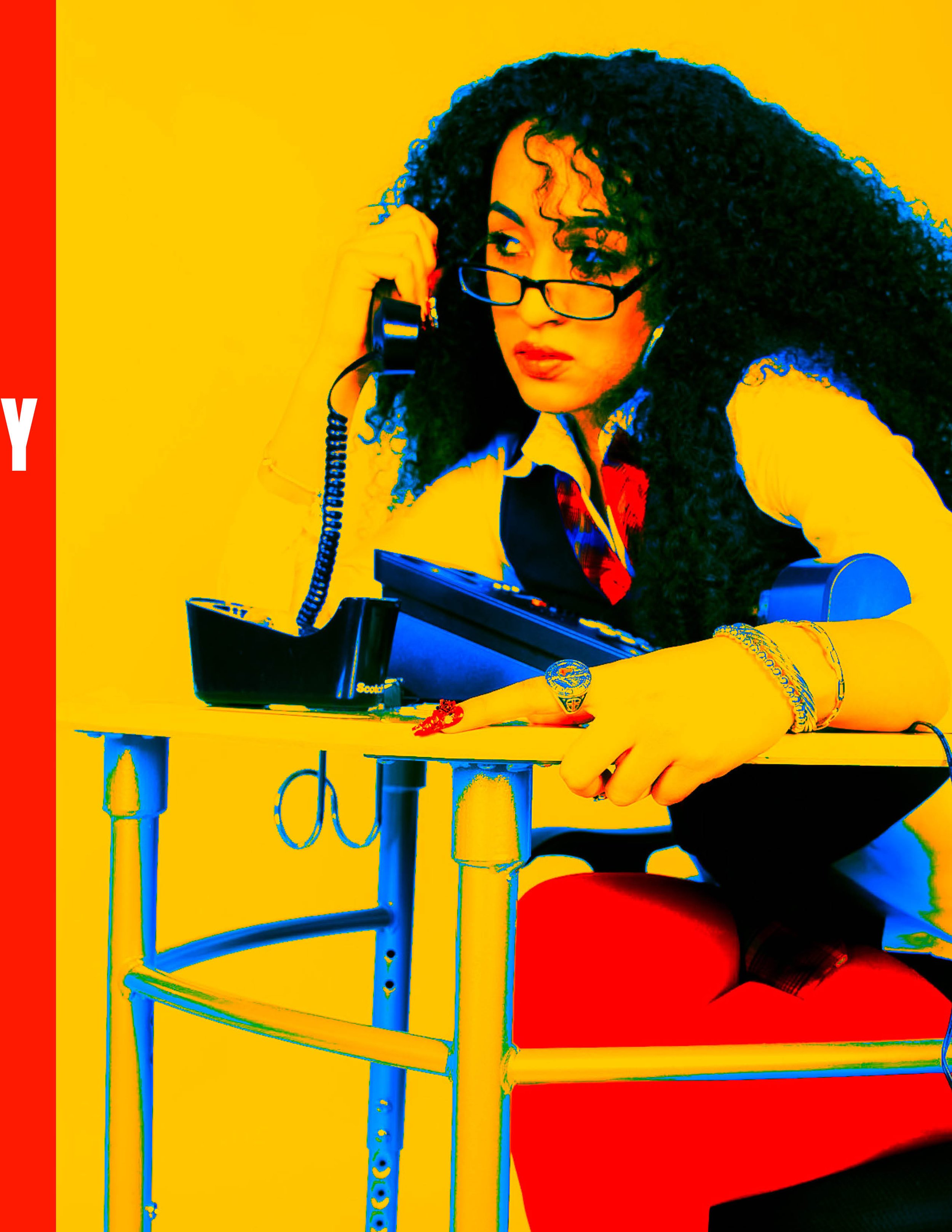 Woman with curly hair and glasses sitting at a desk with a telephone, holding the receiver to her ear, against a yellow background.