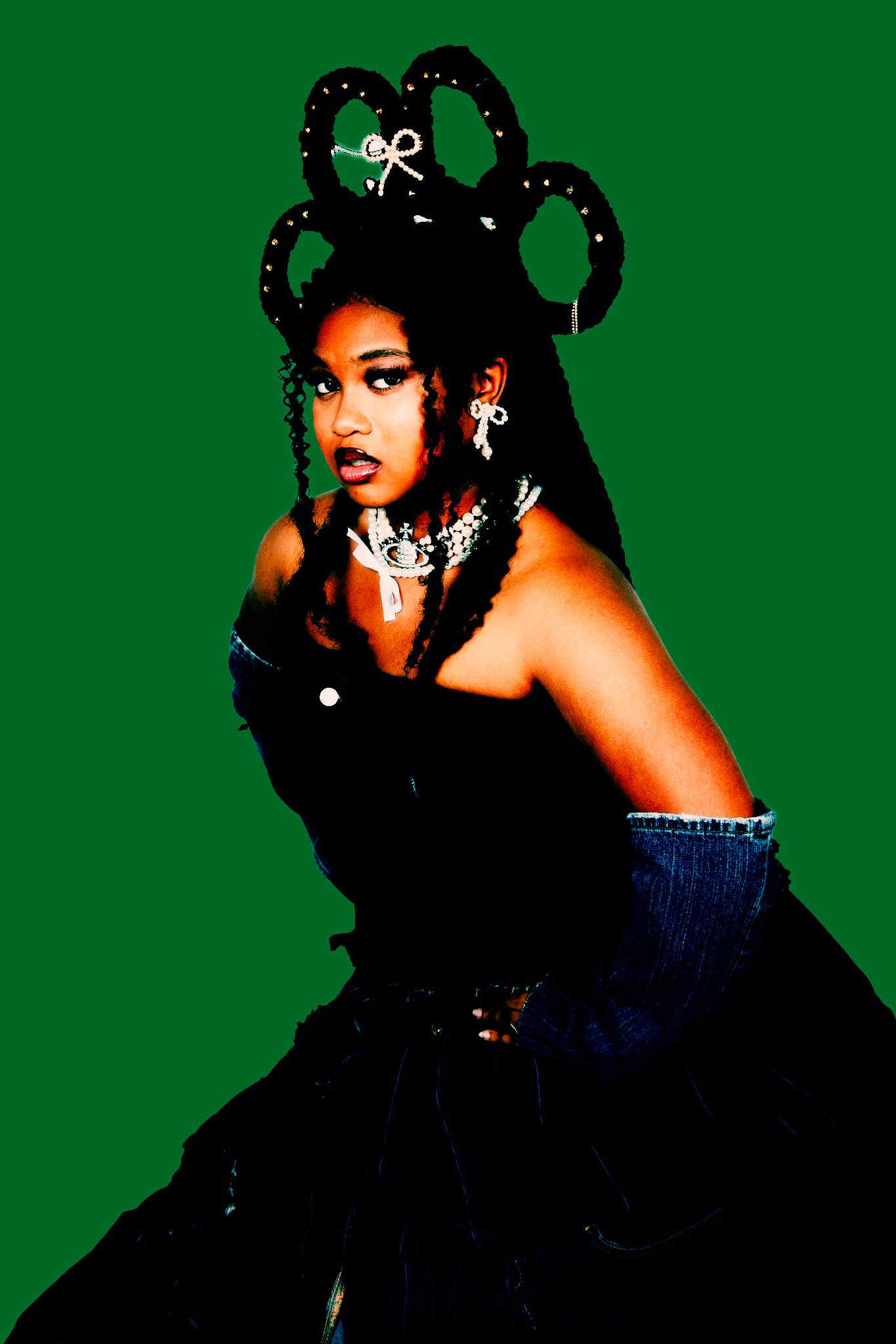 A woman with dark, curly hair styled into large, elaborate loops, wearing jewelry including earrings, necklaces, and a bracelet. She is dressed in a sleeveless top and denim jeans, posing against a solid green background.