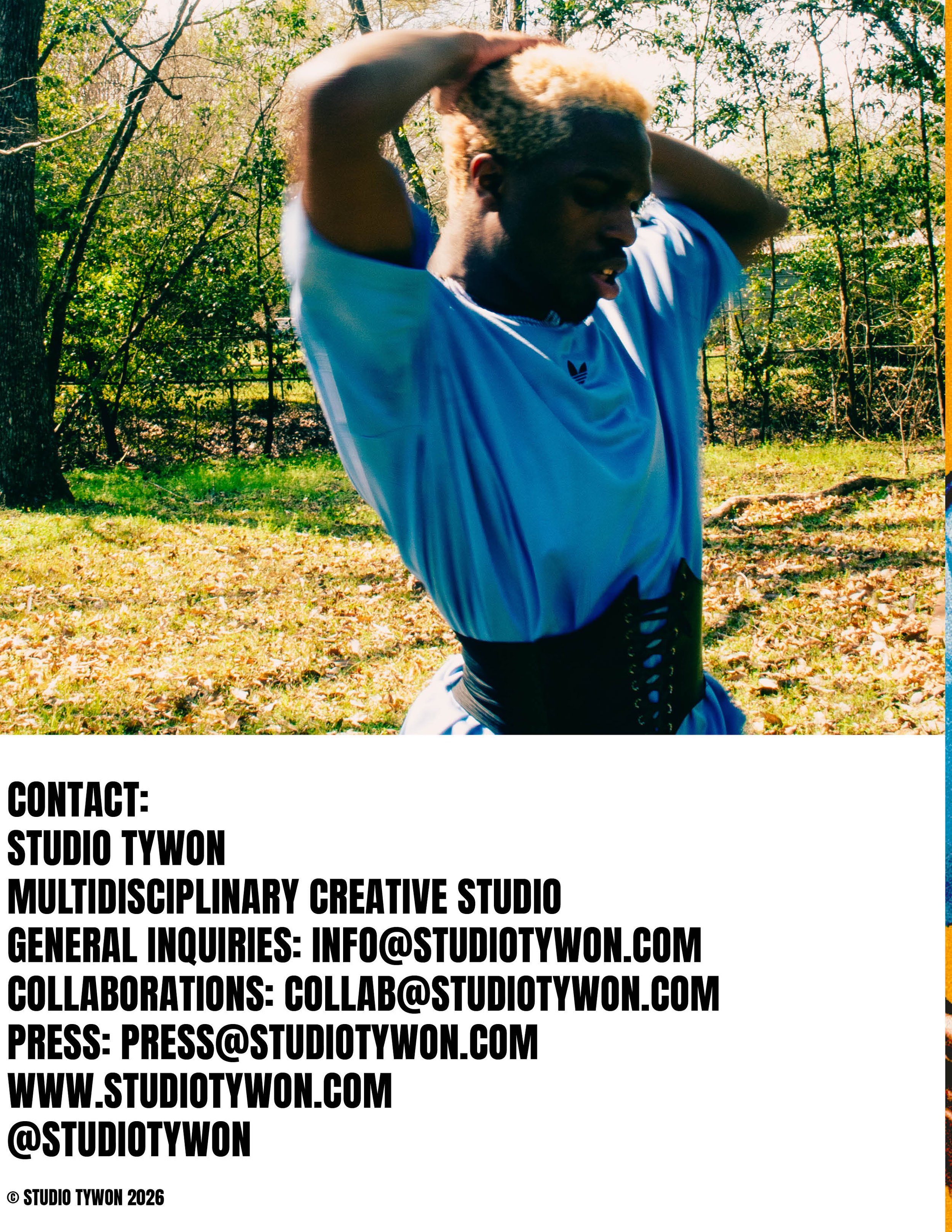 A person with short, blonde hair wearing a light blue t-shirt and a waist trainer is outdoors in a wooded area with green trees and fallen leaves on the ground, adjusting their hair.