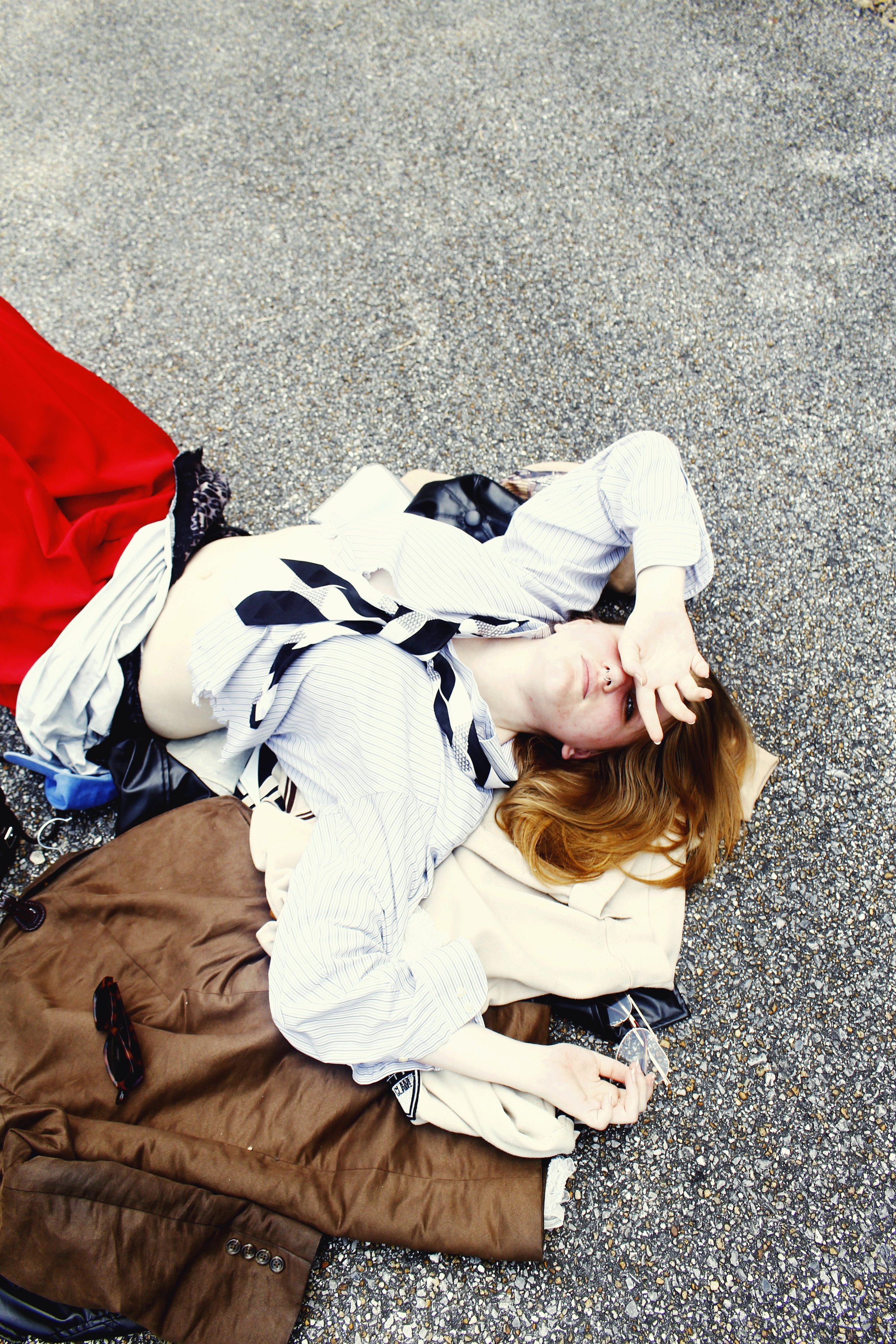 A person lying on the ground on their side, with their head resting on a beige pillow. They are wearing a striped shirt, a black and white scarf, and sunglasses, holding onto a small object in their hand. They are surrounded by various clothing items