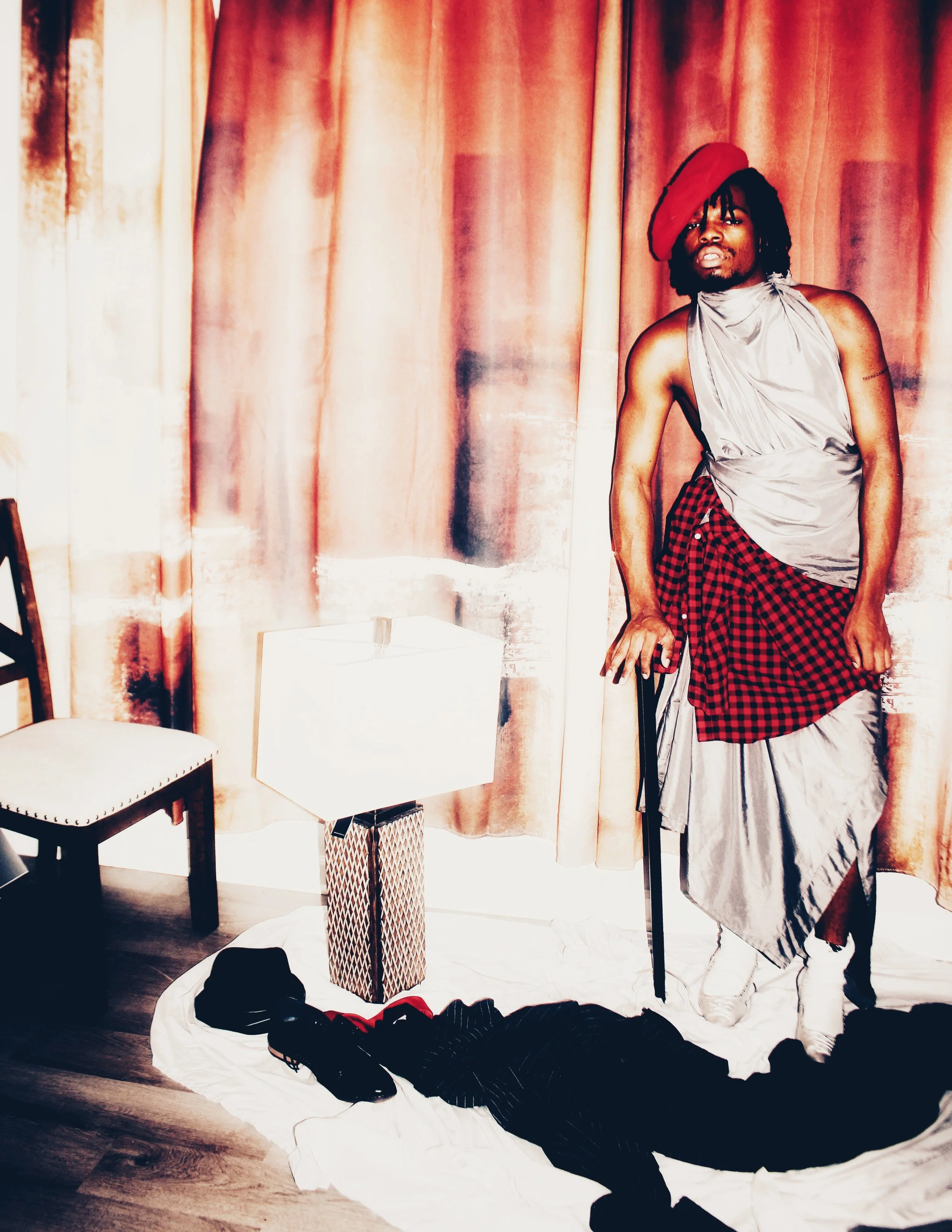 A person wearing a red beret, grey sleeveless top, red and black checkered skirt, and white shoes, standing beside a chair, a white lampshade, and some clothing items on the floor in front of a pink curtain.