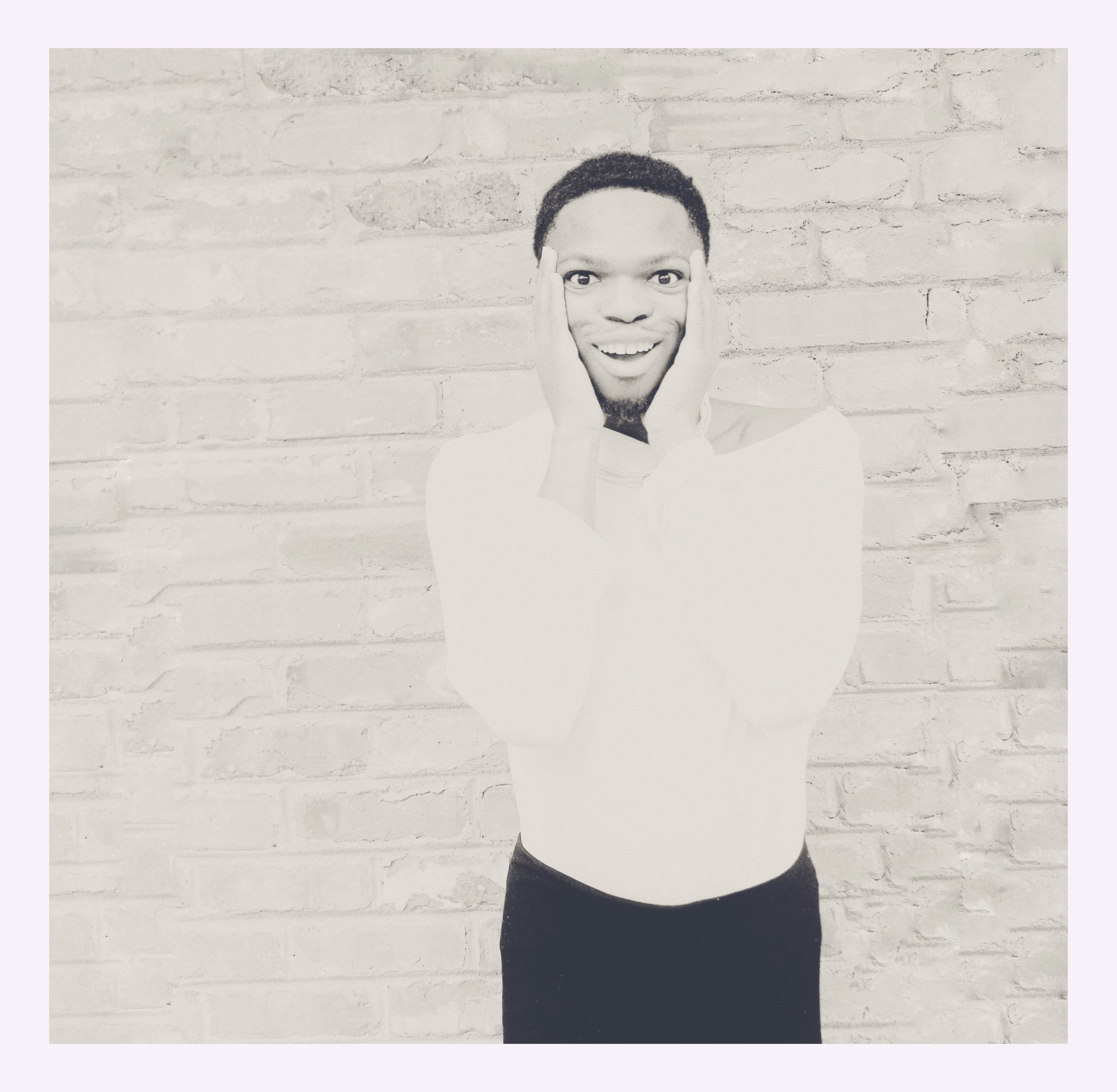 Black and white photo of a surprised person with hands on face, smiling, standing against a brick wall.