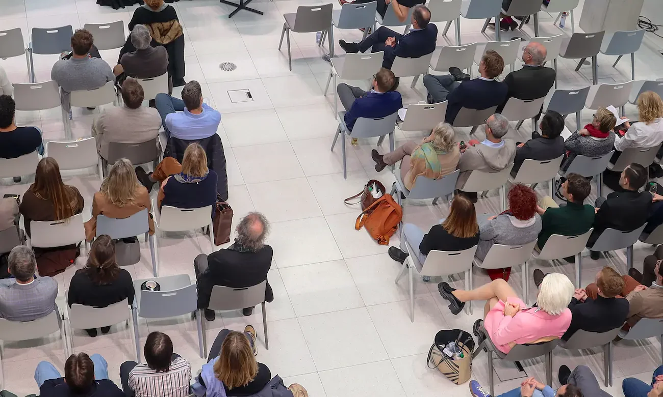 Group of people sitting in front of a stage, everyone wears dark clothes. Betty Business wears a pink dress and stands out.