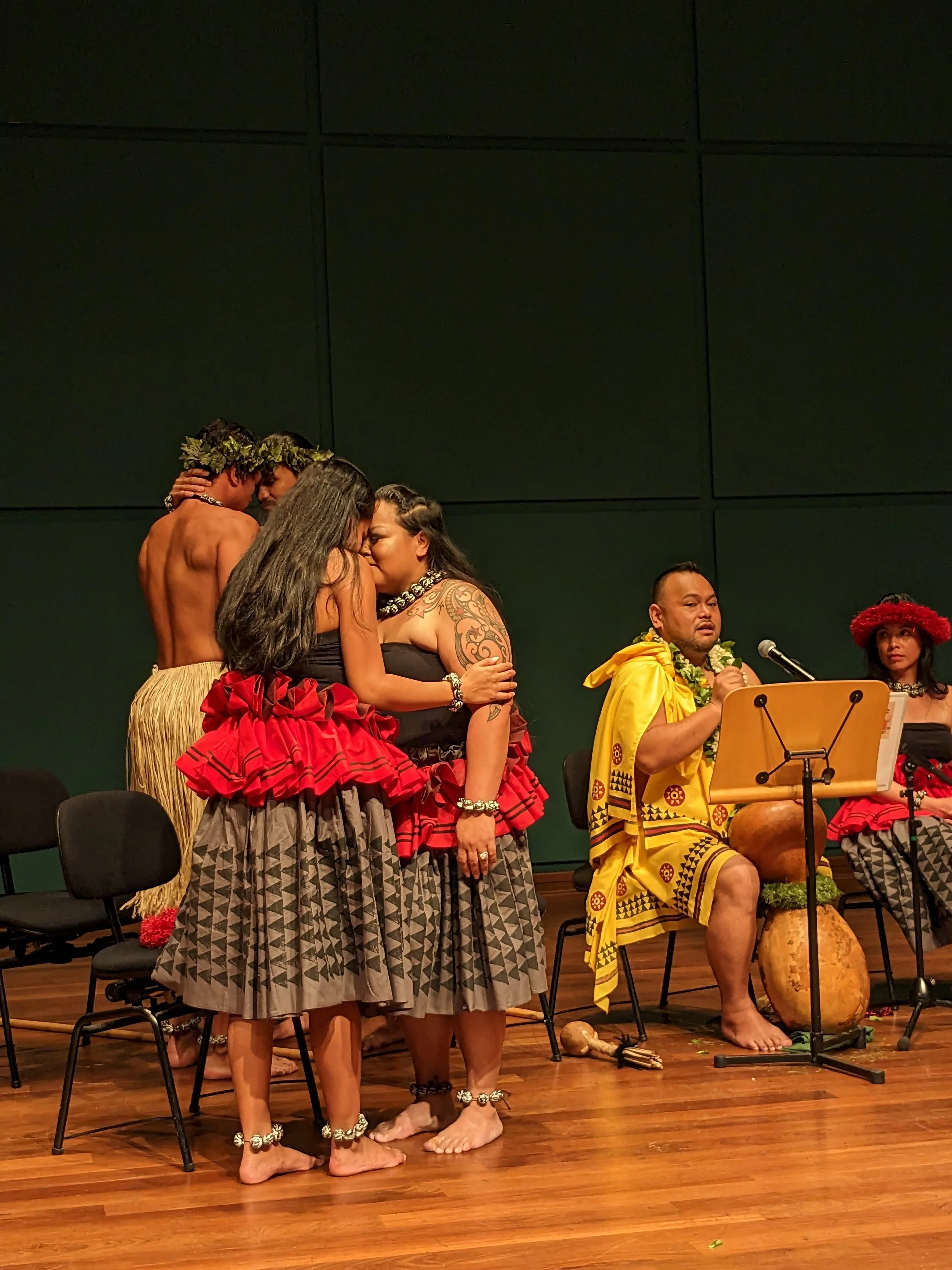 Hawaiian ceremony with two women kissing, dressed in traditional attire, and seated individuals wearing Hawaiian clothing playing musical instruments.