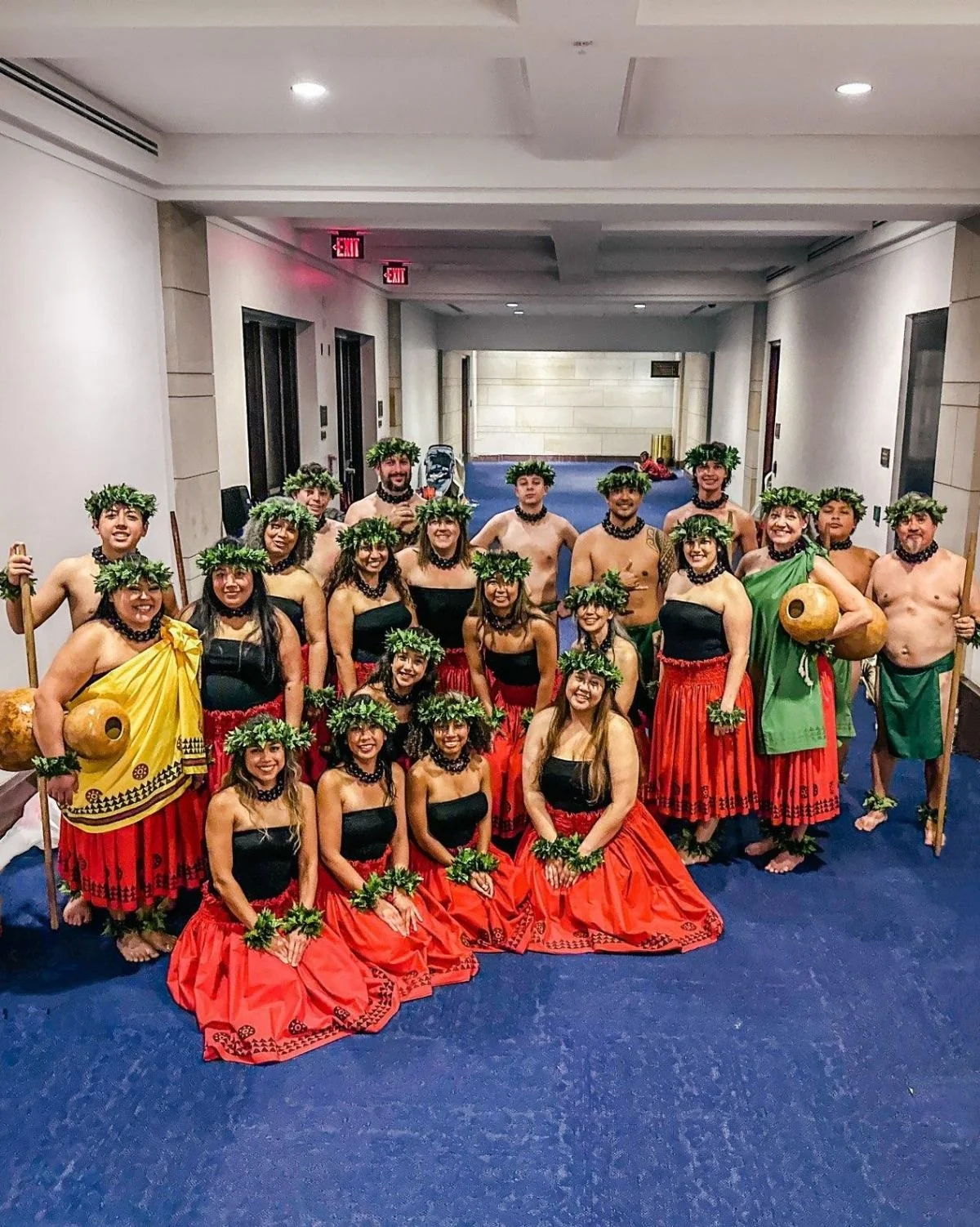 Group of people in traditional Polynesian costumes, wearing floral crowns, black tops, and red skirts, standing on a blue carpet in an indoor hallway.