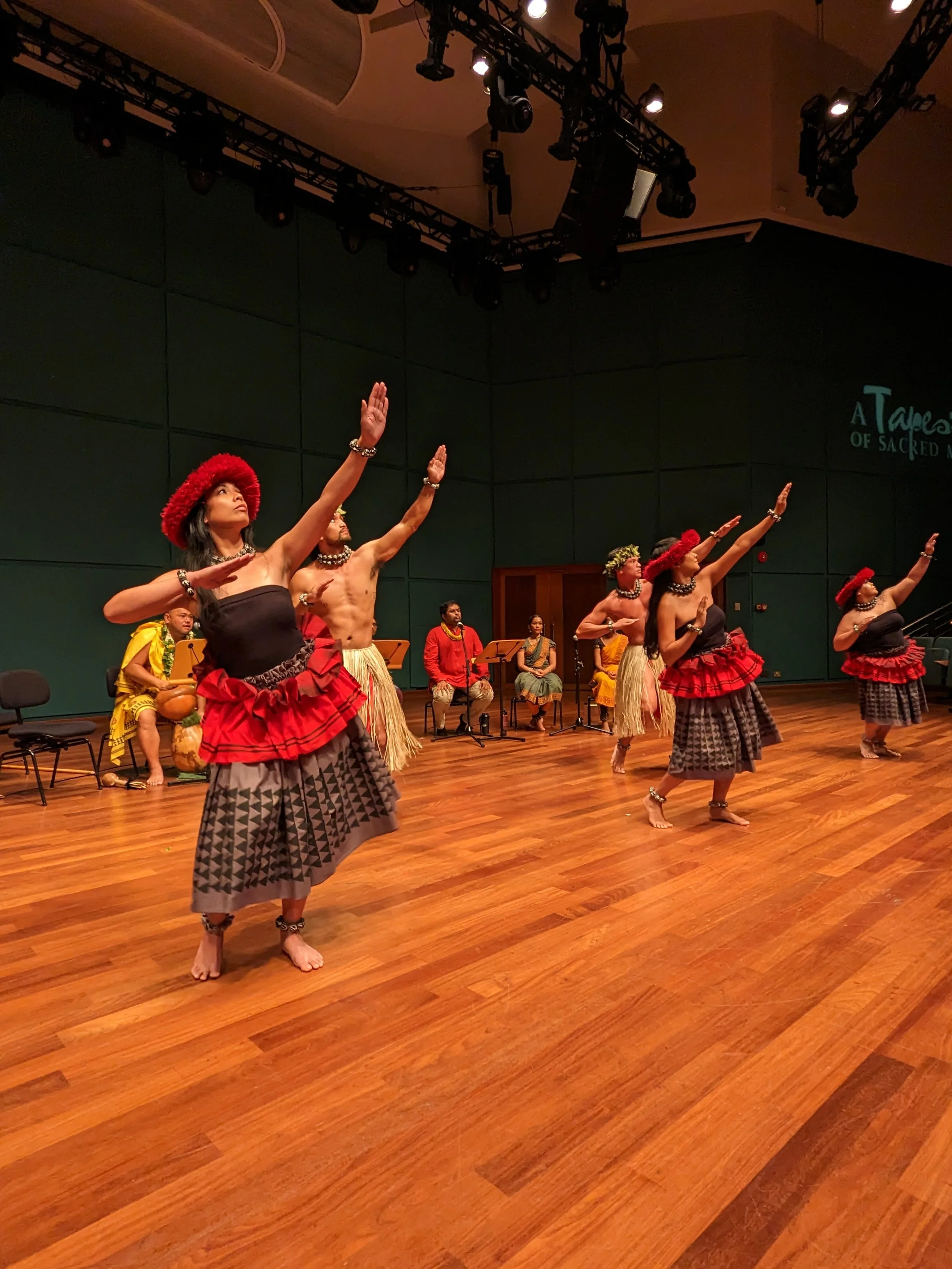 Traditional Hawaiian hula dancers performing on stage with four women in front wearing grass skirts and red headbands, and four men behind in yellow, red, and traditional attire, seated and playing musical instruments.