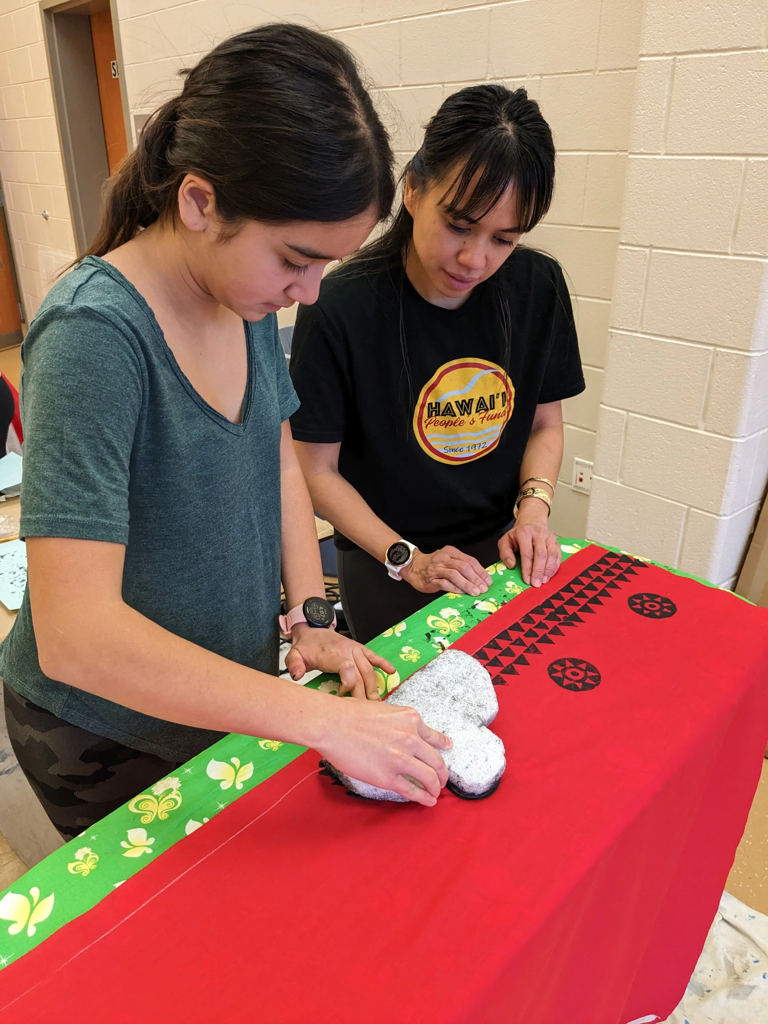 Two women are working on a craft project with a red and green tablecloth. One woman is pressing a glittery Mickey Mouse-shaped object onto the table. They are focused on their task in a room with beige brick walls.