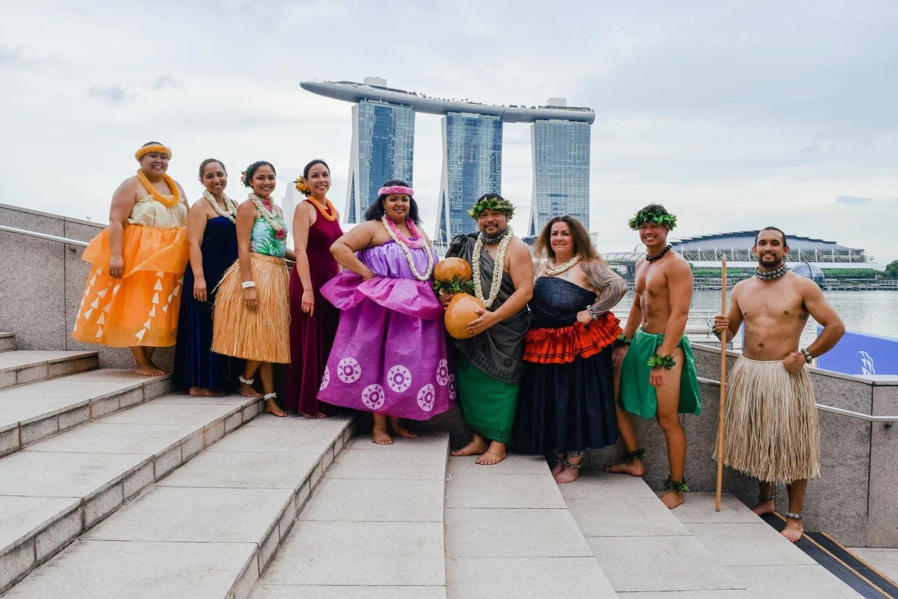 Group of people in traditional Polynesian attire standing outdoors with Marina Bay Sands hotel in the background.