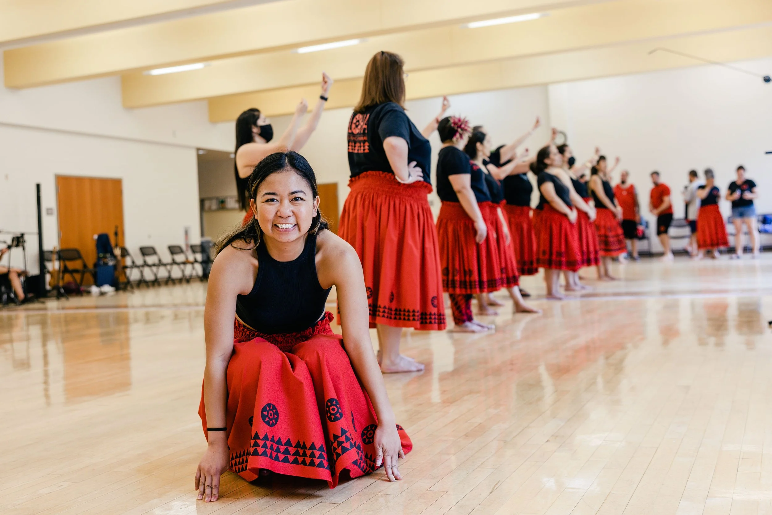 A group of women in traditional red skirts and black tops practicing hula dancing in a dance studio, with one woman smiling and kneeling in the foreground.