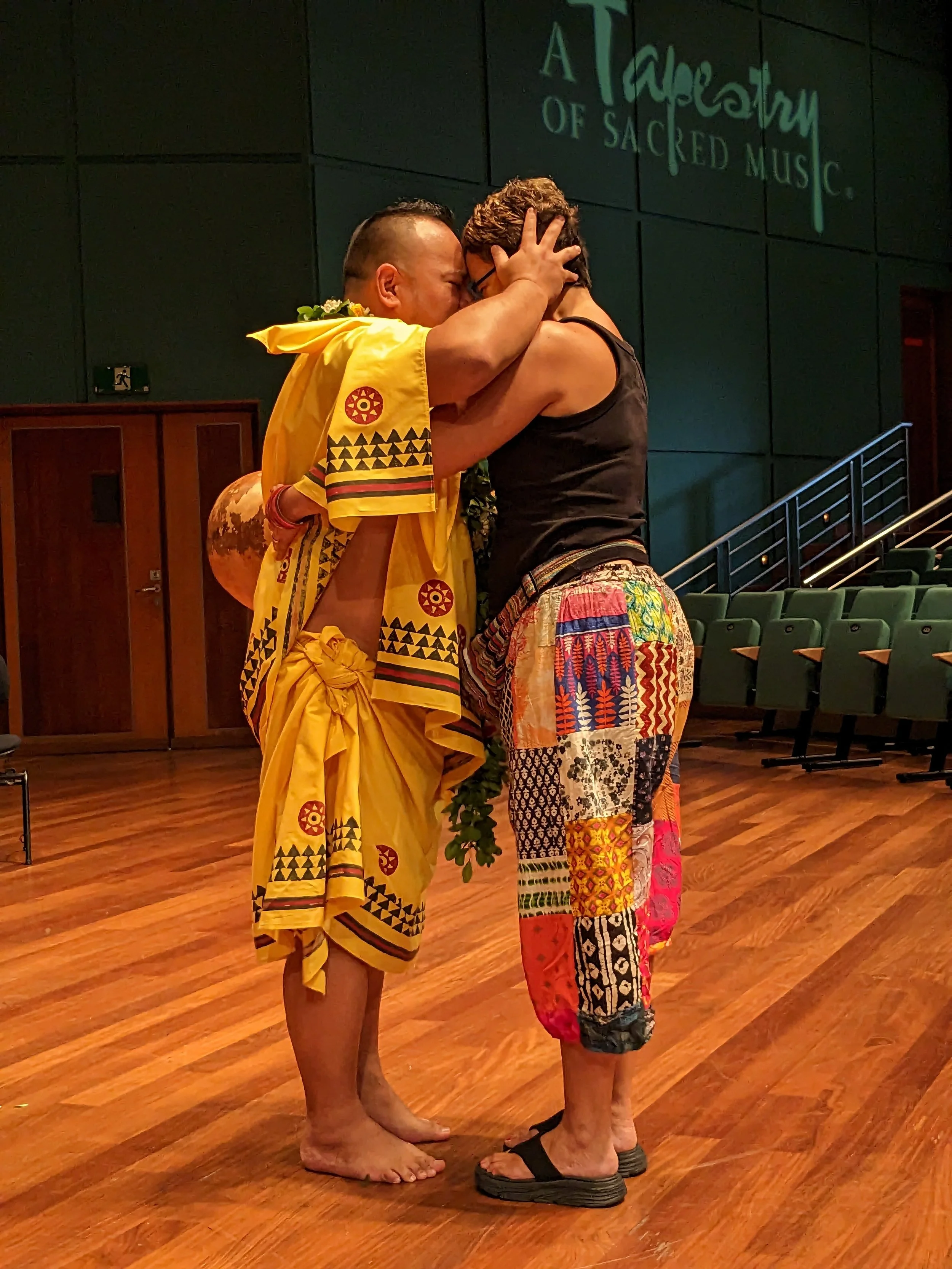 Two people are embracing in a dance or ceremony on a wooden stage; one person is dressed in traditional yellow Polynesian attire with tribal patterns, and the other is wearing colorful patterned pants and a black tank top.